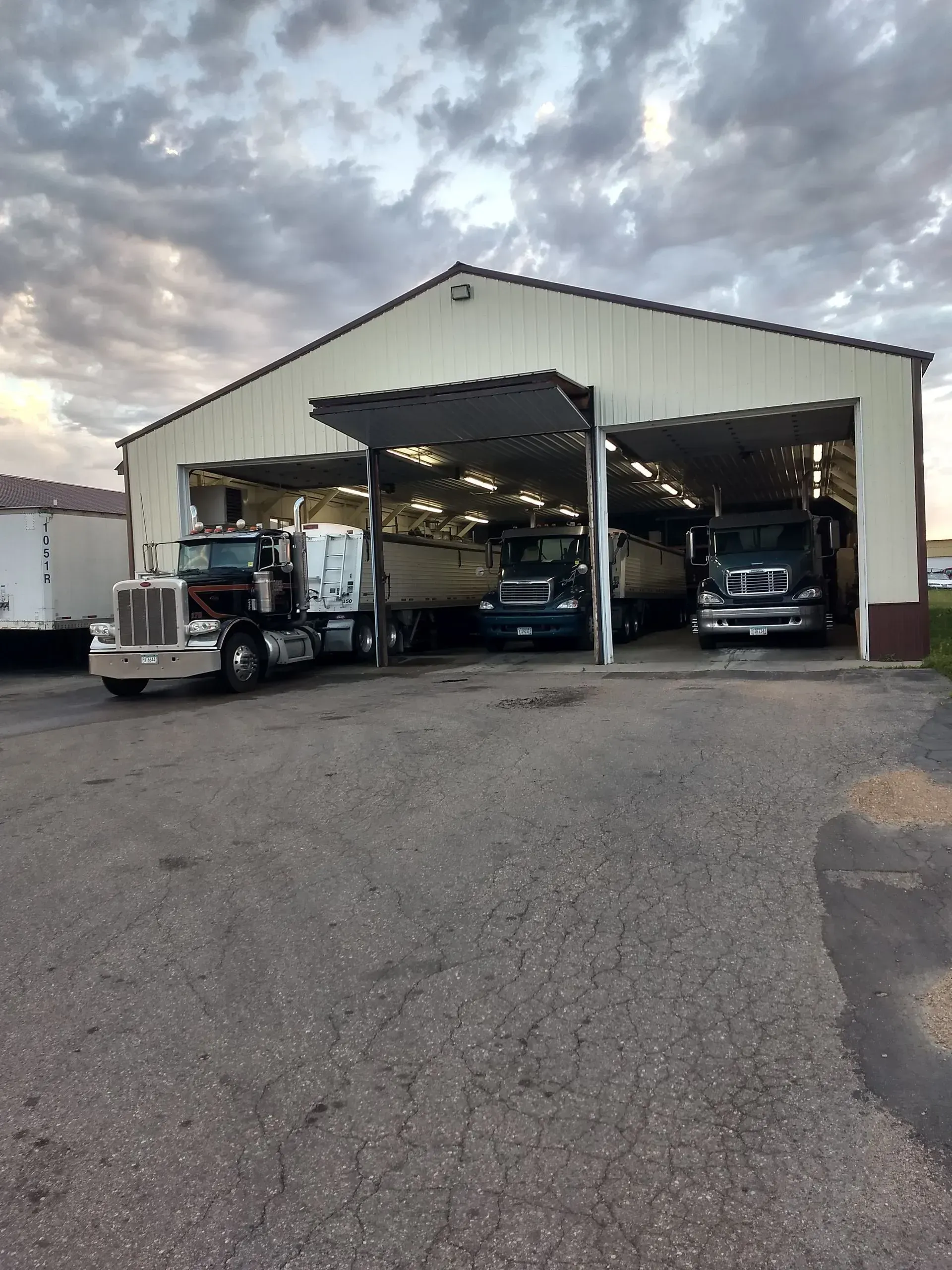 Three semi-trucks parked inside a large, open-bay garage at dusk under a cloudy sky.