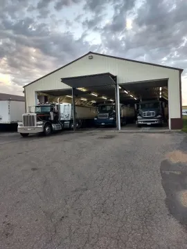 Three semi-trucks parked inside a large, open-bay garage at dusk under a cloudy sky.
