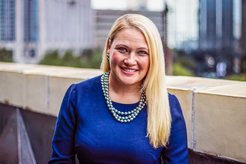 Smiling woman in a blue top and beaded necklace standing by a concrete railing outdoors