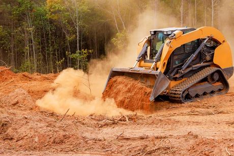 Orange skid-steer loader digging dirt in a muddy forest clearing, kicking up dust