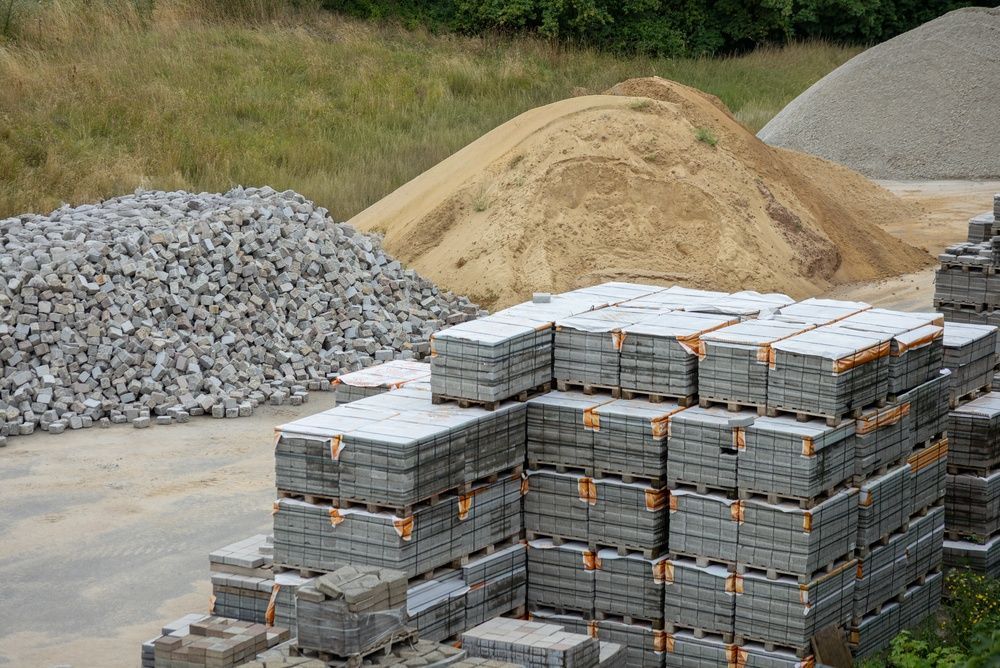 Stacks of gray paving stones beside gravel and sand piles at a строитель materials yard