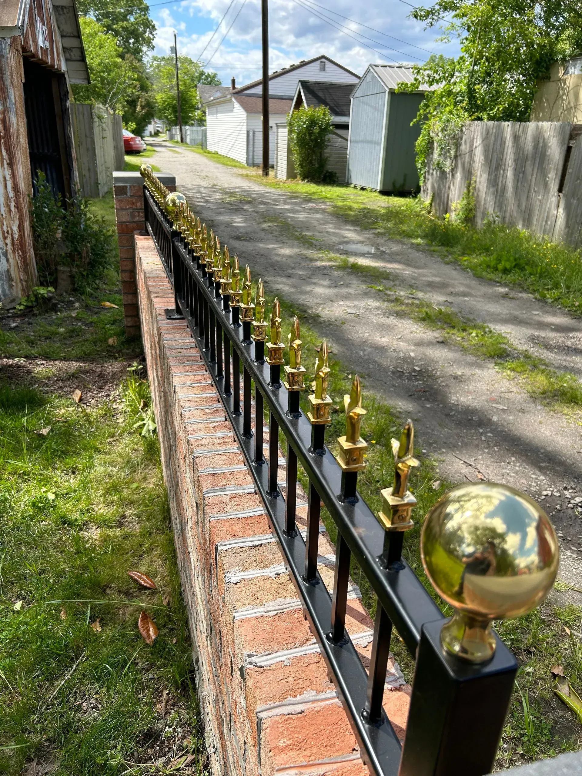 Brick wall topped with a black metal fence featuring gold finials, bordering a gravel alleyway.