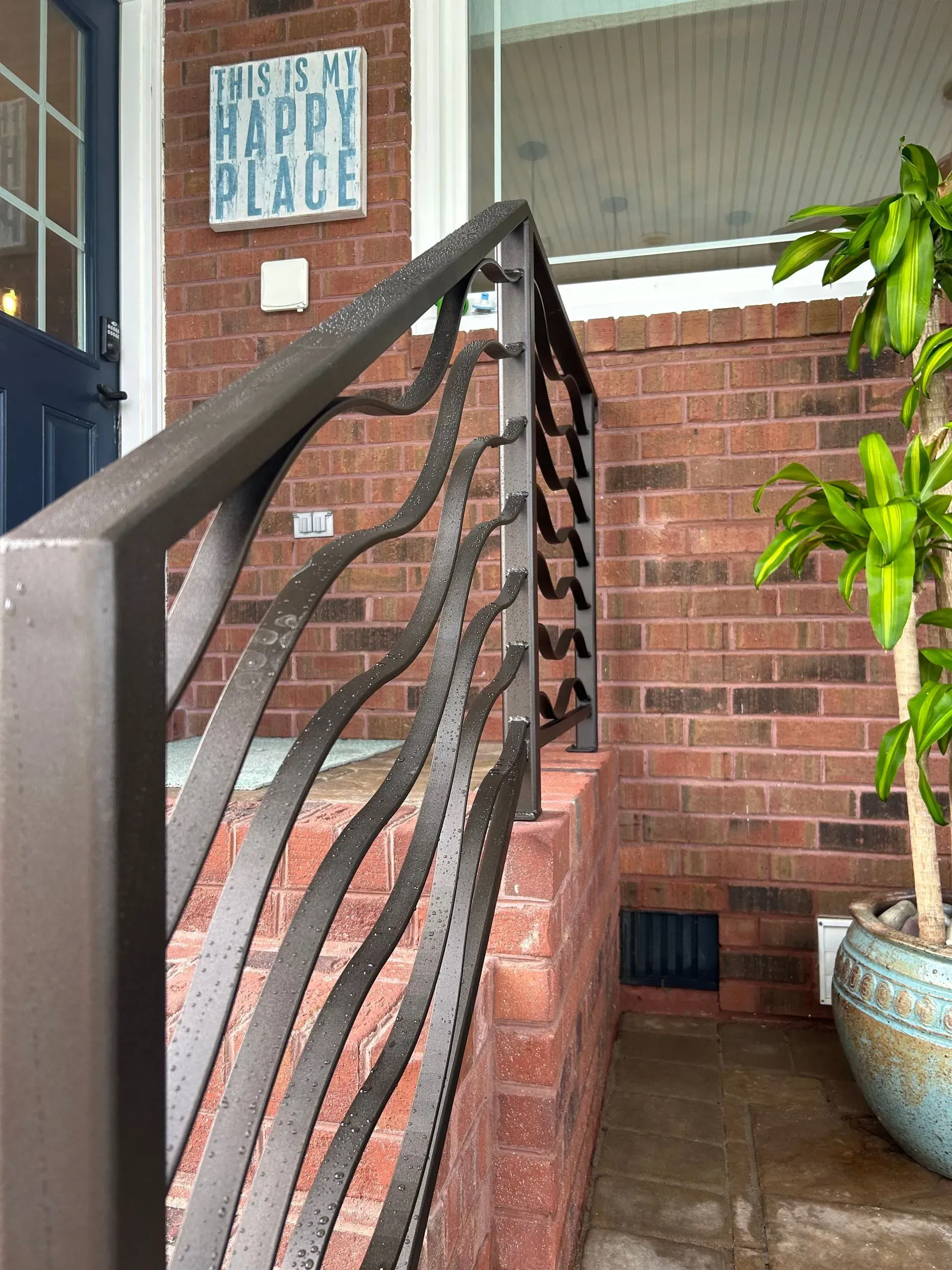 Metal railing with a wavy design on a brick porch; a potted plant sits to the right.