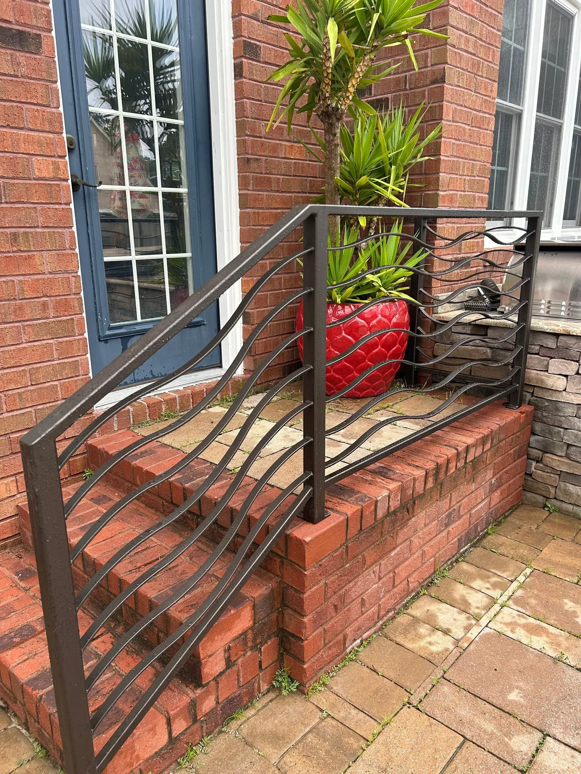 Brown metal handrail with a wavy design on brick steps, next to a red planter and a blue door.