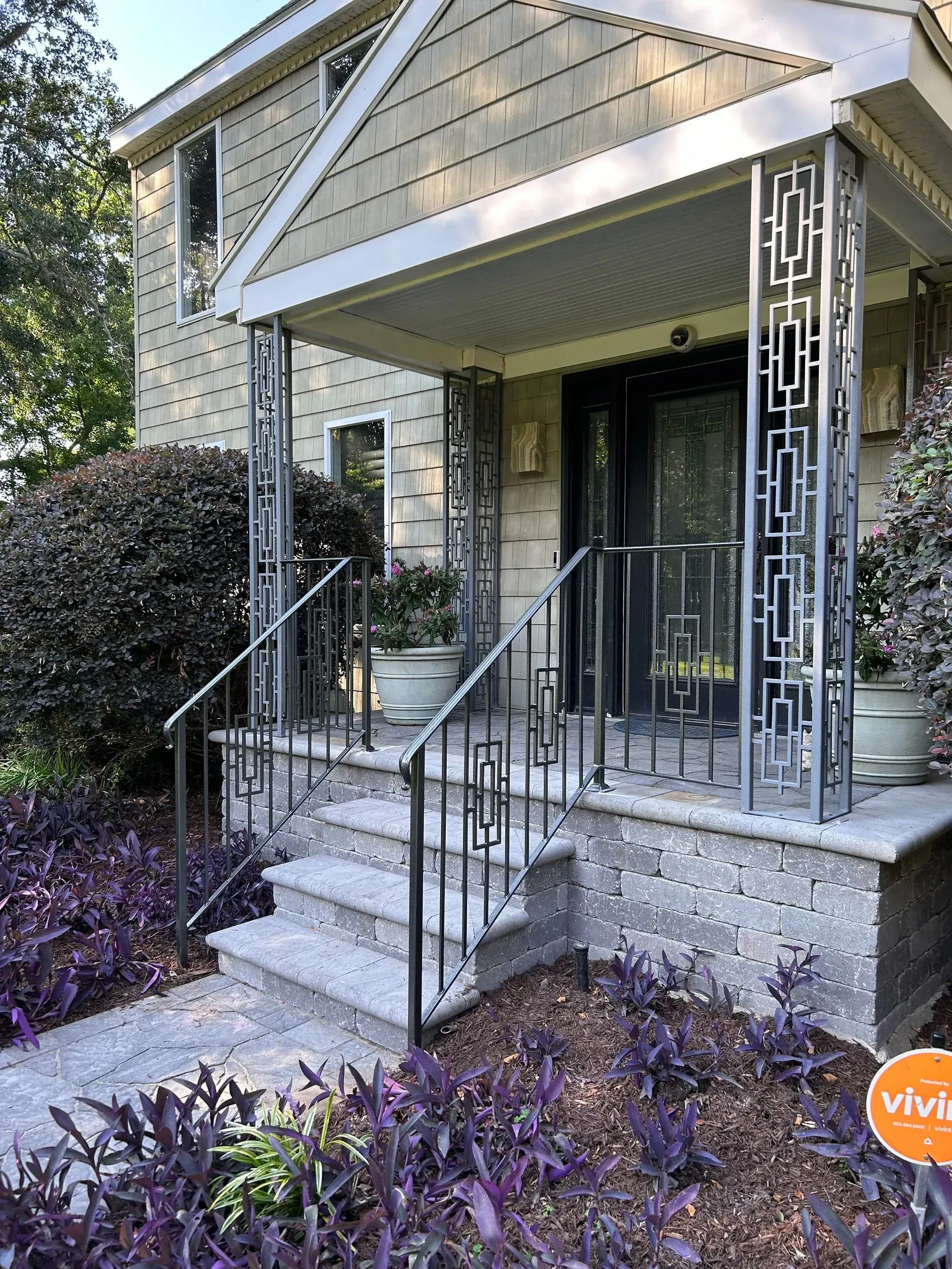 Front porch of a house with decorative metal railings and steps. Purple ground cover and a gray stone facade.