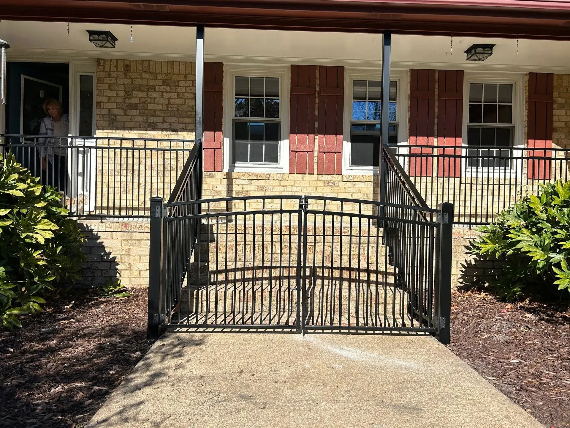 Black metal gate and railings leading to a brick house with red shutters. Person stands in the doorway.