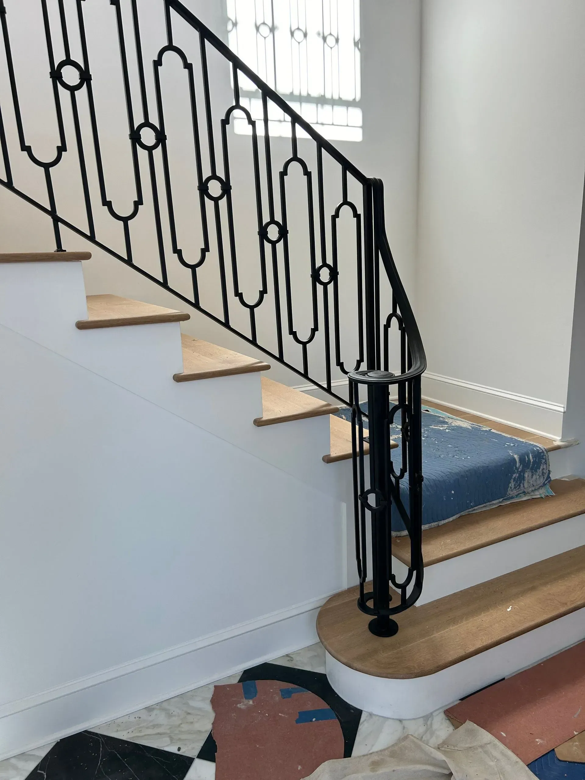 Black wrought-iron railing on a staircase with wooden steps and white walls. A patterned rug is visible on the floor.