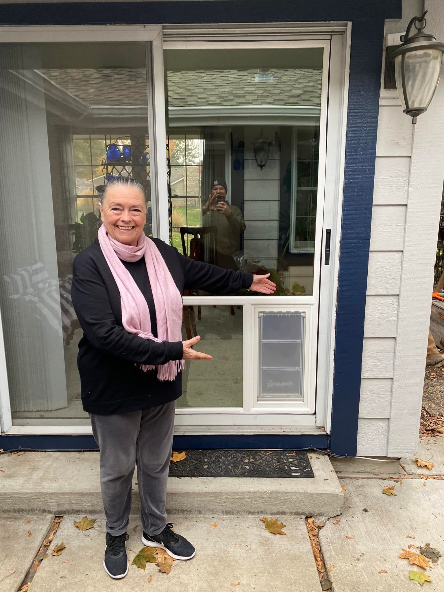 Woman points to a pet door installed in a window. She's outside a house, wearing a scarf.