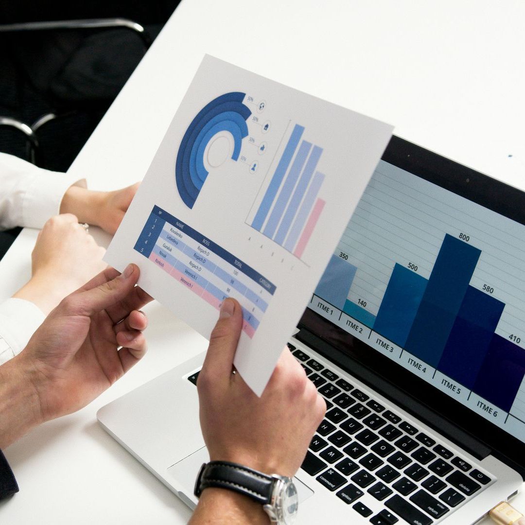 Hands holding and pointing to a chart with bar and pie graphs, next to a laptop displaying similar data.