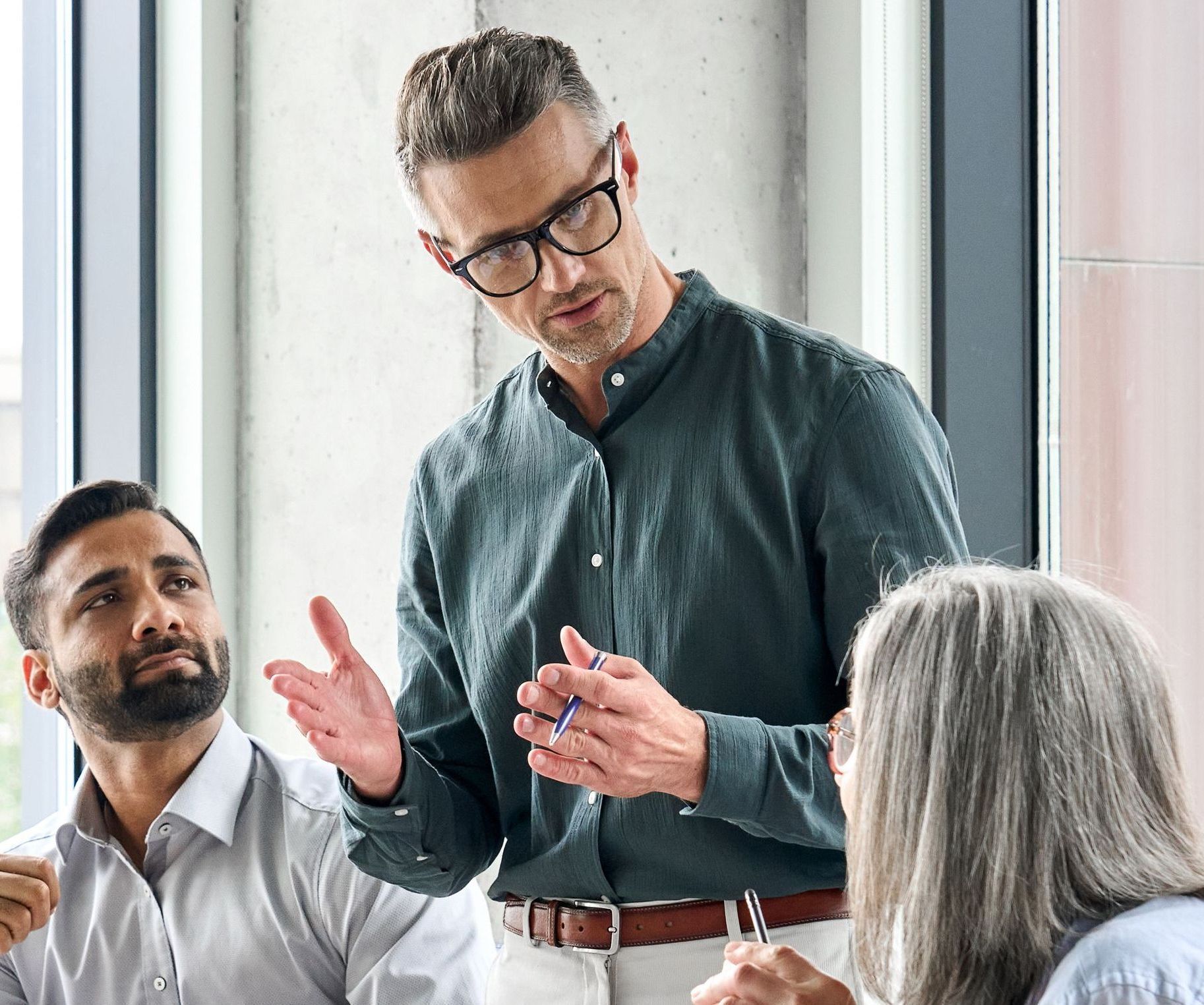 Man speaking in a meeting, gesturing with hands; two colleagues listen.