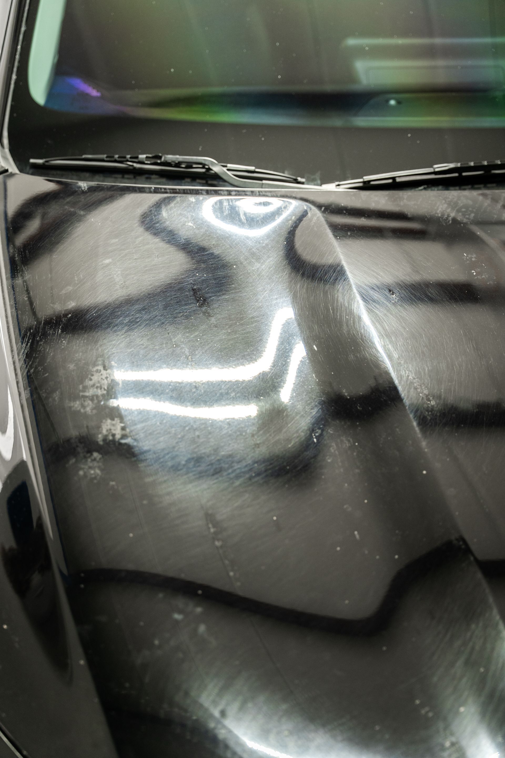 Close-up of a glossy black car hood with bright overhead reflections and a windshield at the top