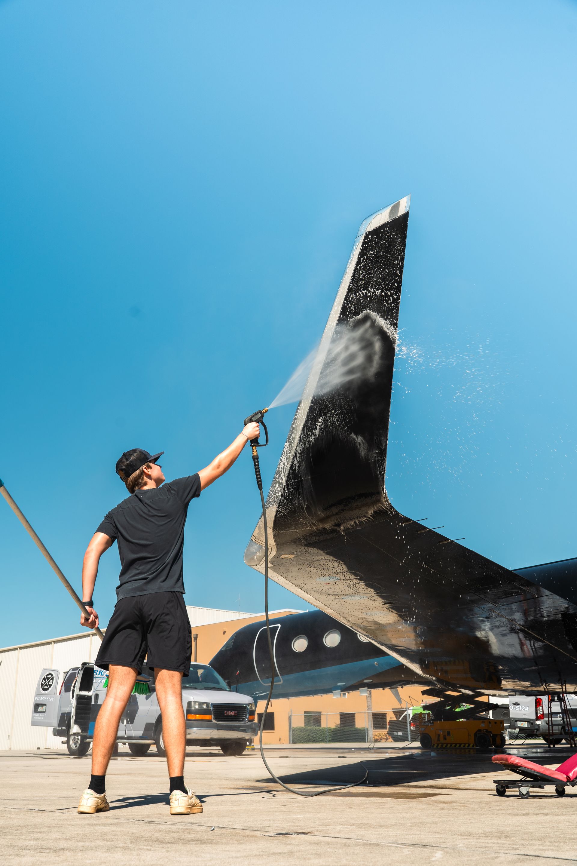 Person spraying water on a black sail or wing-shaped surface outdoors under a clear blue sky