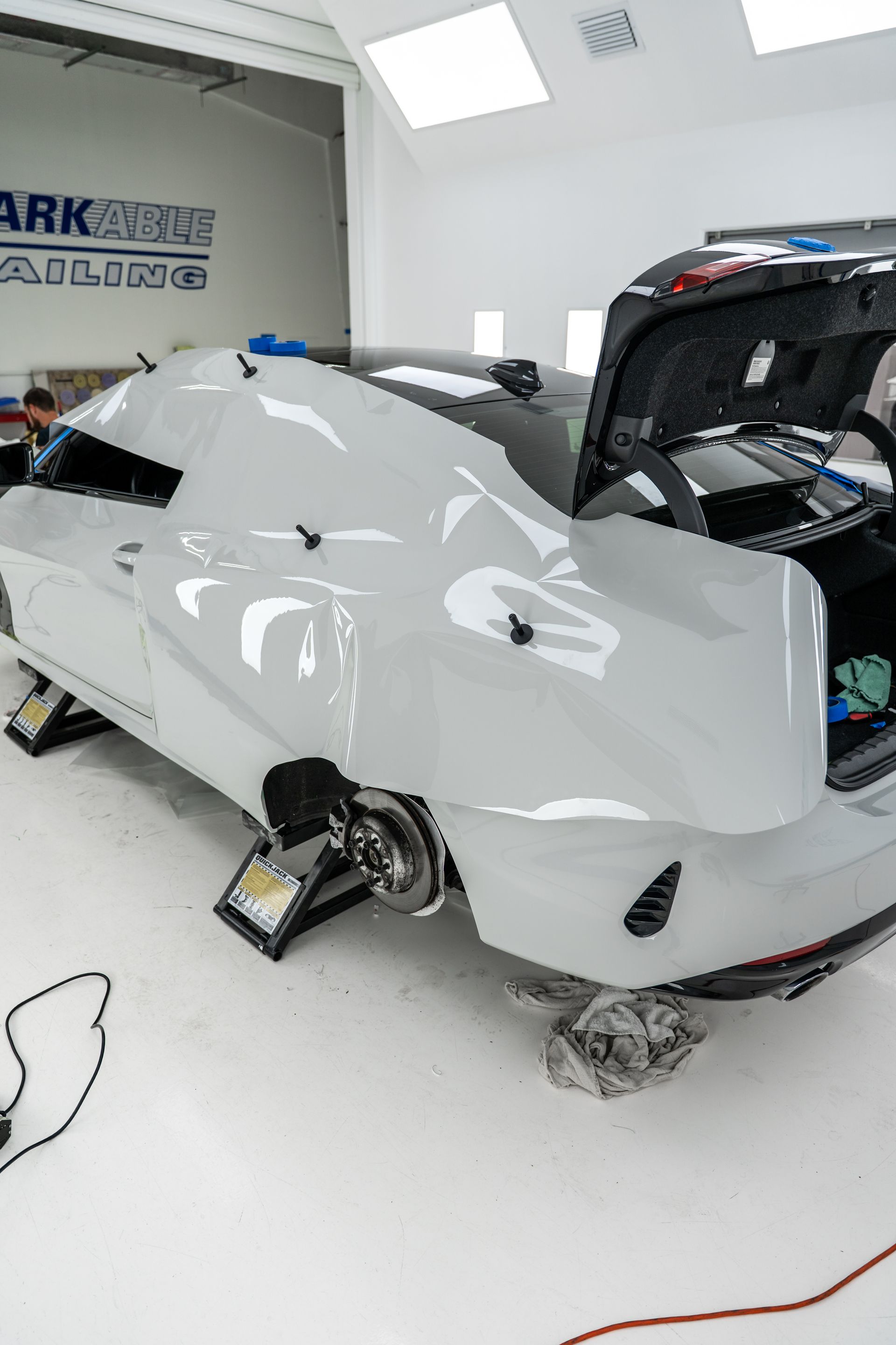 White sports car in a repair shop, raised on jacks with rear bodywork removed.