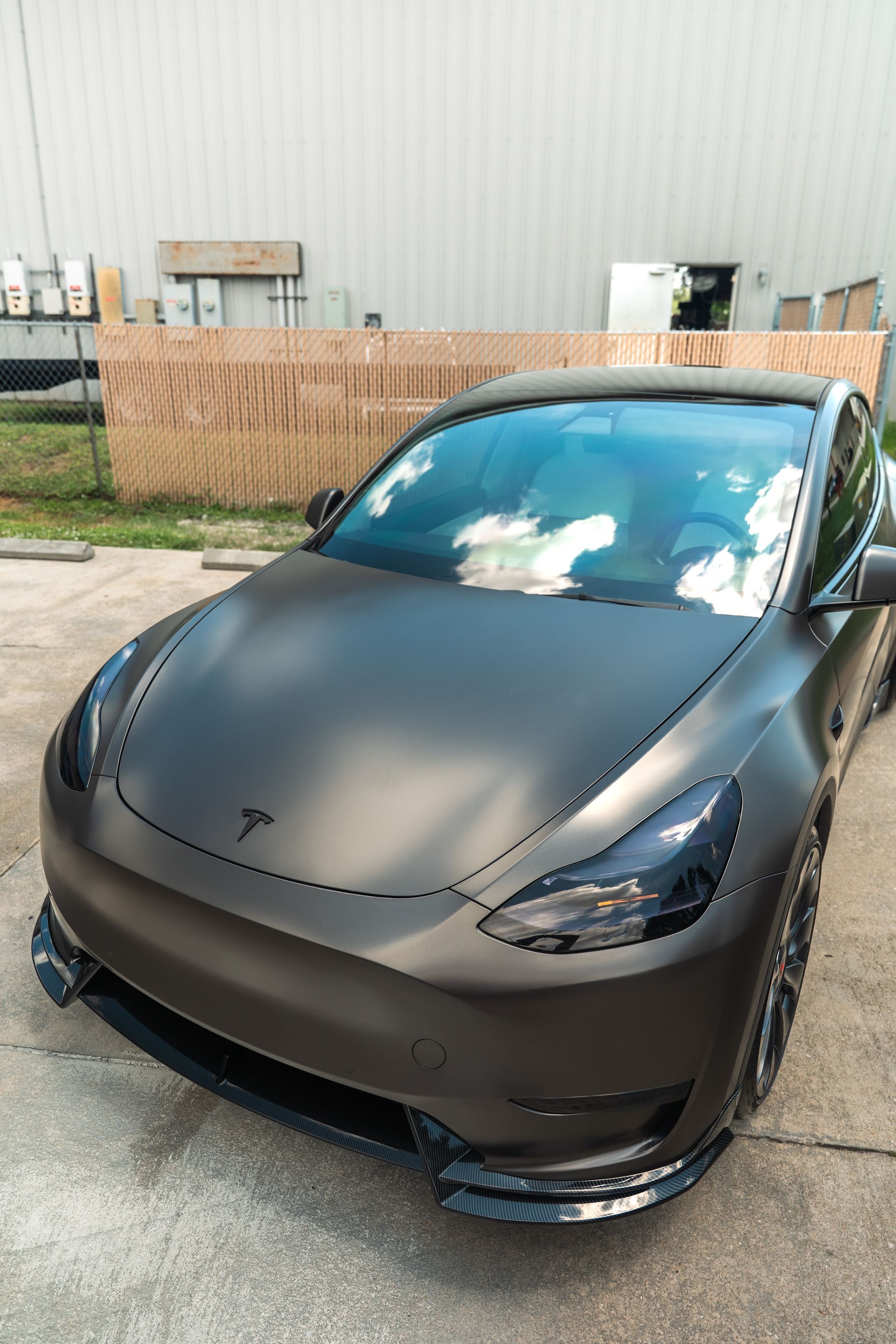 Matte black Tesla parked outdoors with a wooden fence and cloudy reflections on the windshield