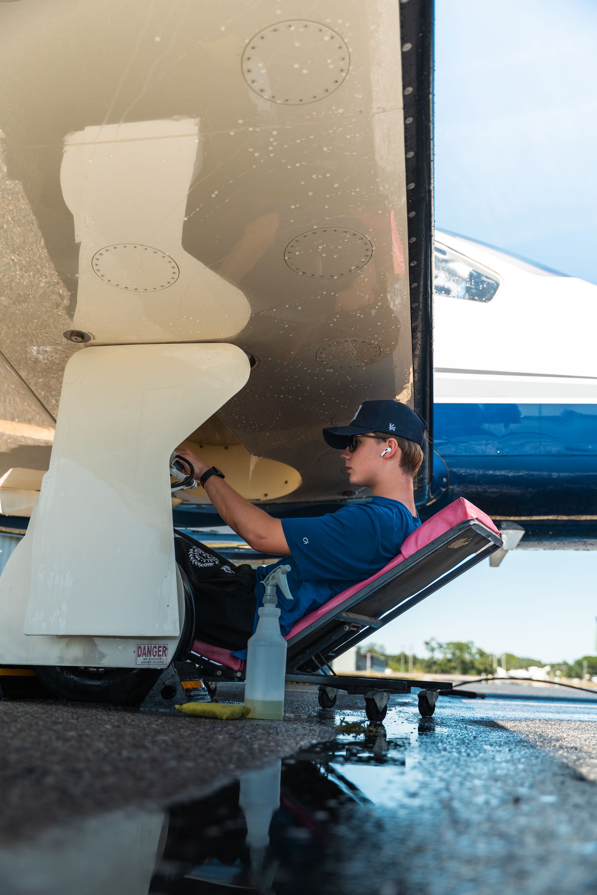 Person seated on a boat painting the hull white with a roller, dock and water visible nearby