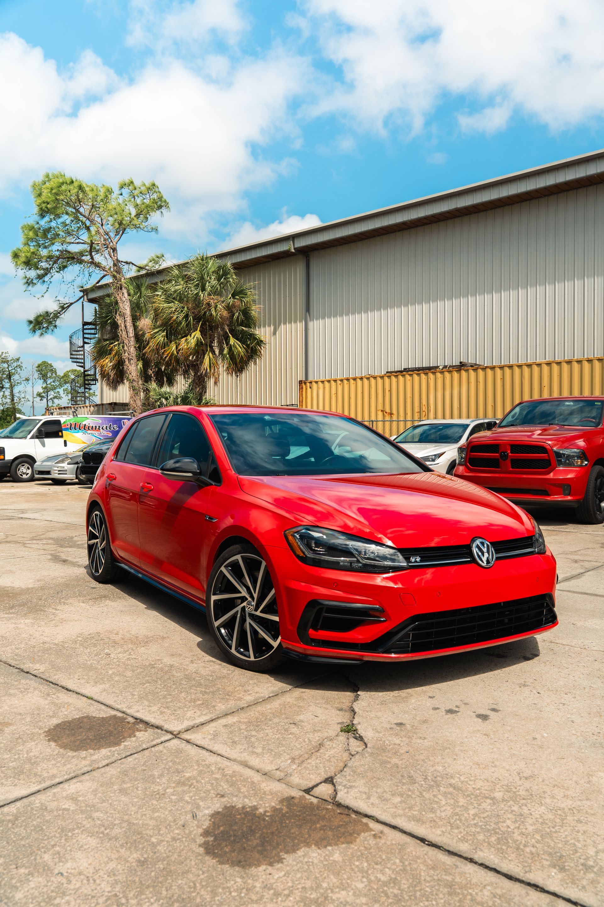Red Volkswagen hatchback parked in front of an industrial building on a sunny day.