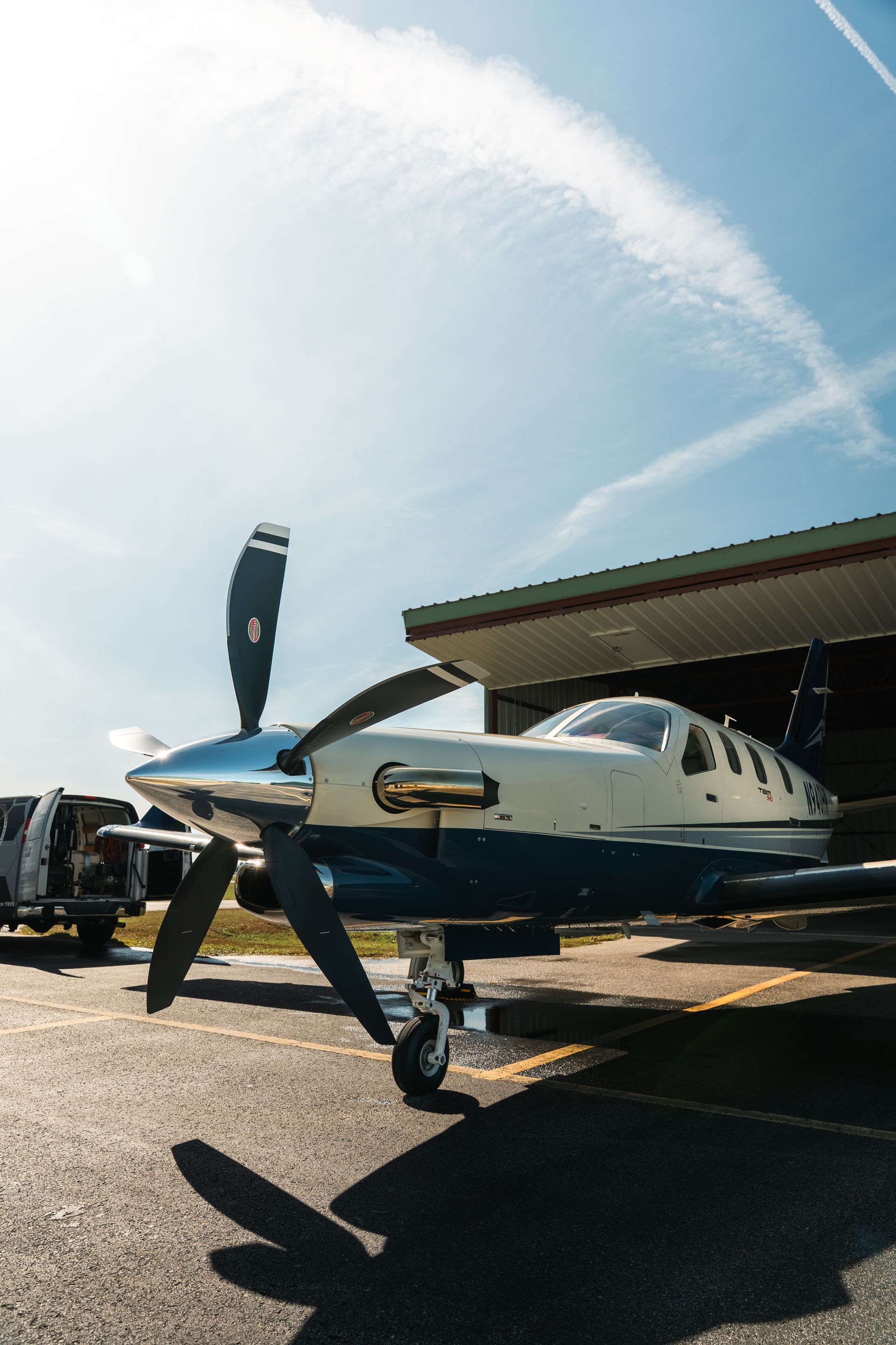 Small blue-and-white propeller airplane parked on a sunny tarmac by a hangar