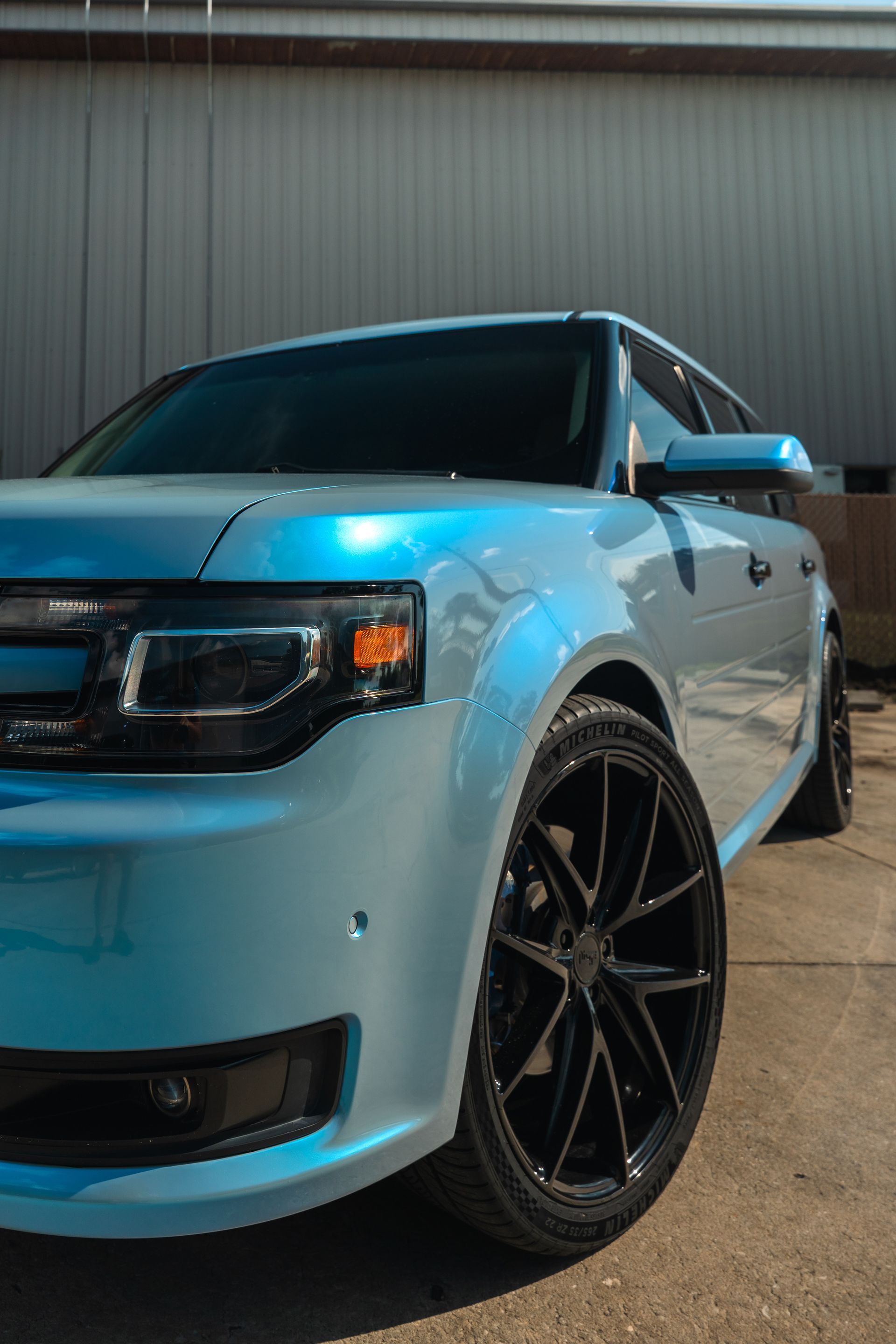 Light blue sports car parked outdoors beside a metal building, shown from the front-left angle.