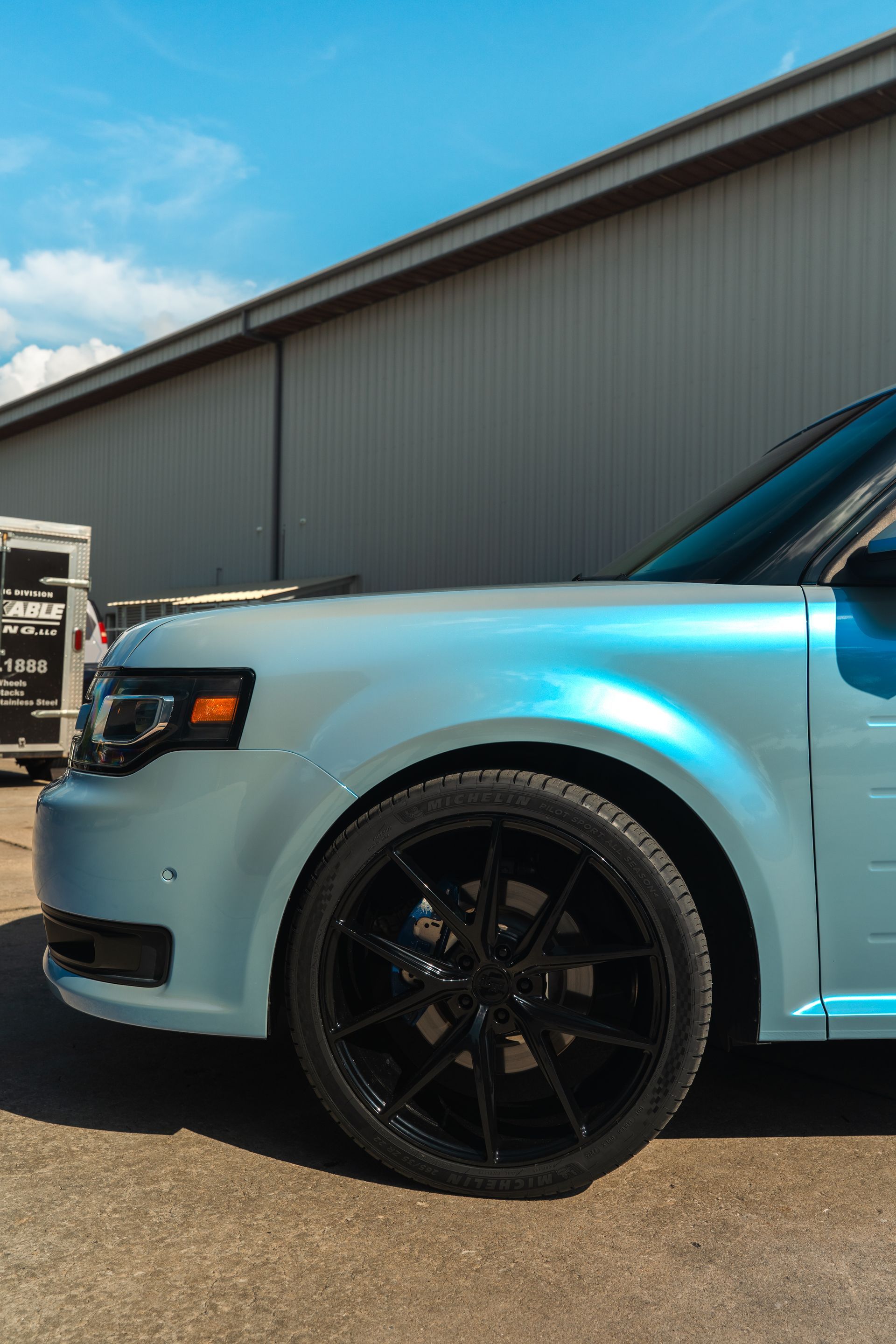 Light blue SUV front side with black wheels parked beside a warehouse under a clear sky