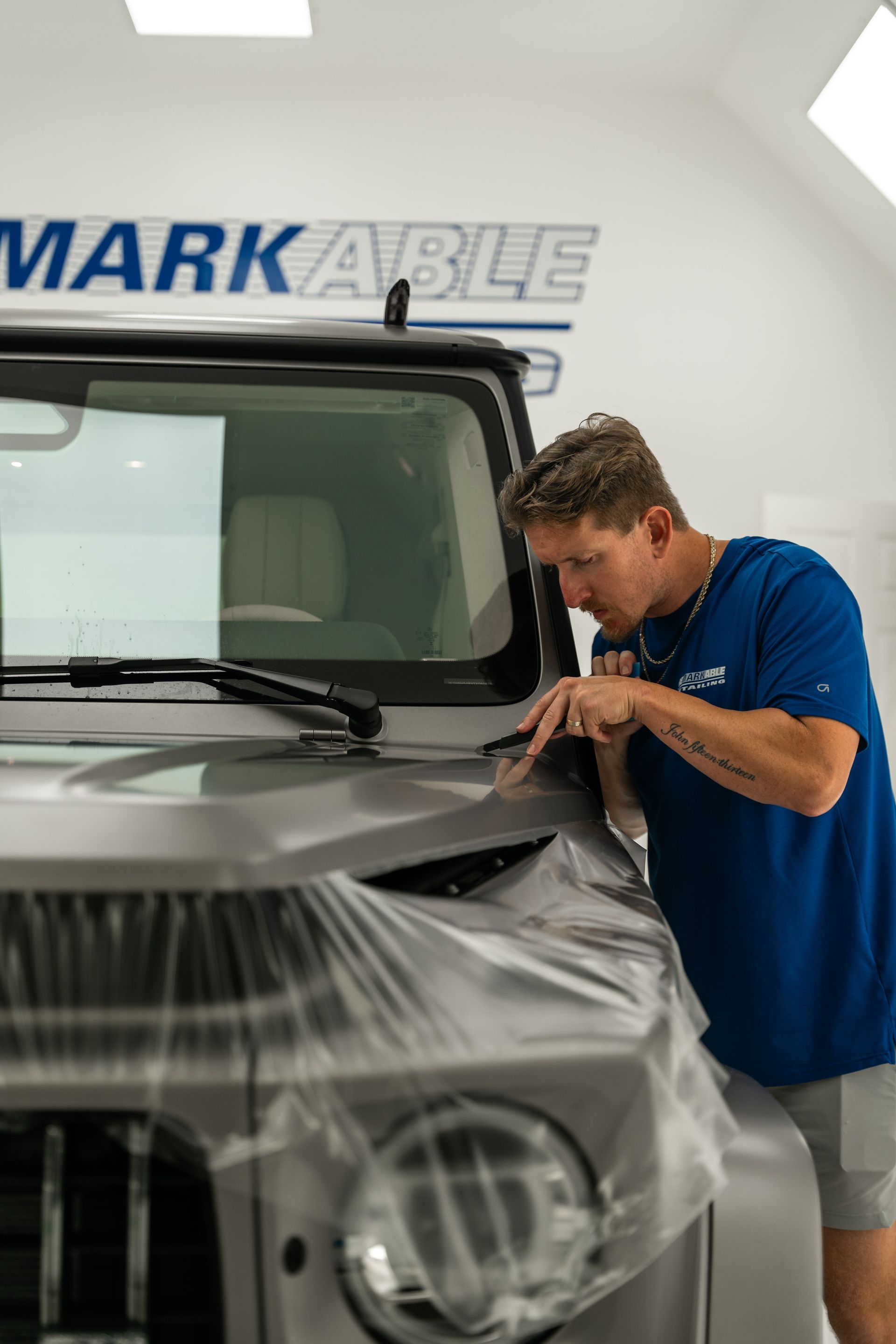 Man in blue shirt inspecting a car hood wrapped in protective plastic inside a detailing shop