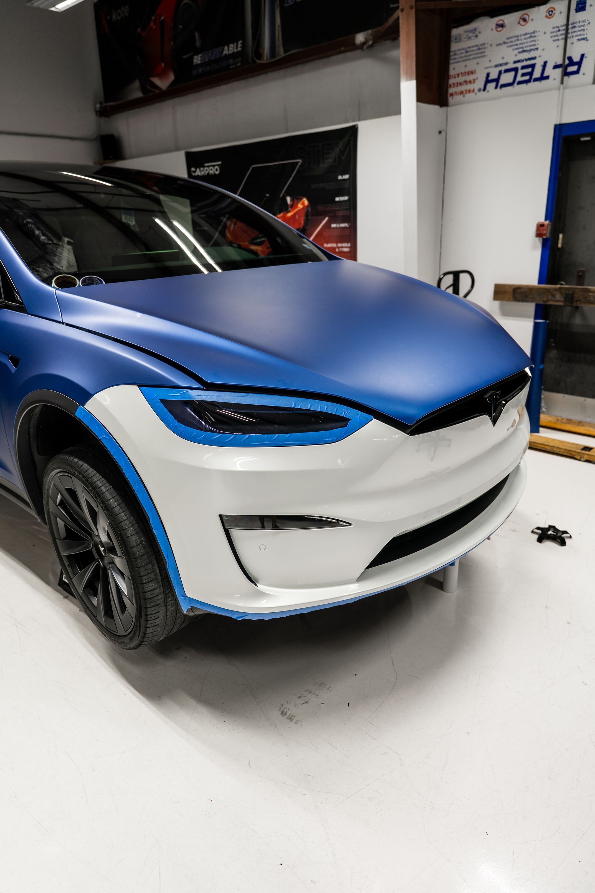 Blue-and-white car in a garage with its front hood open and damaged bumper