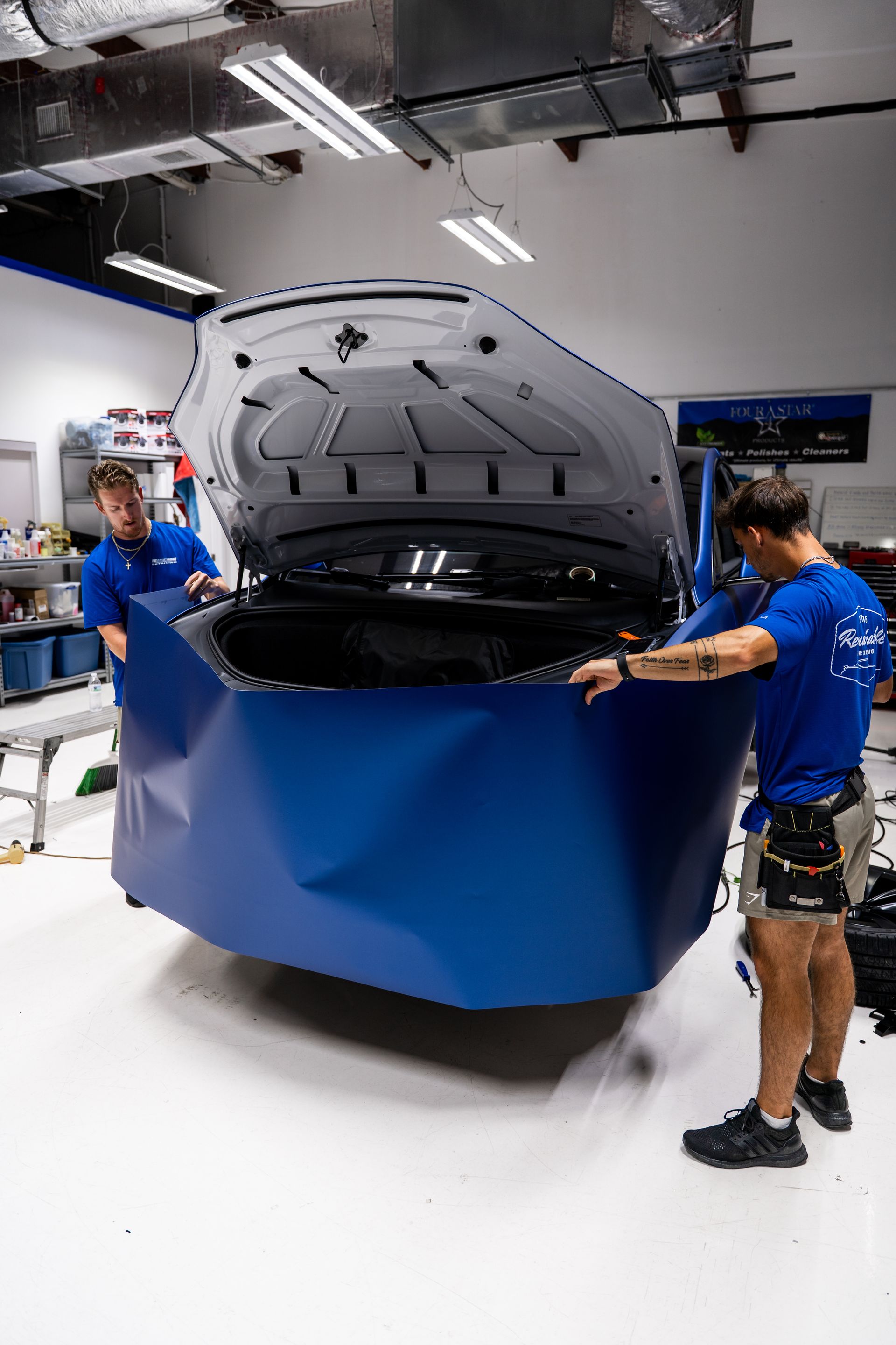 Two workers in blue uniforms open a large car hood in a workshop
