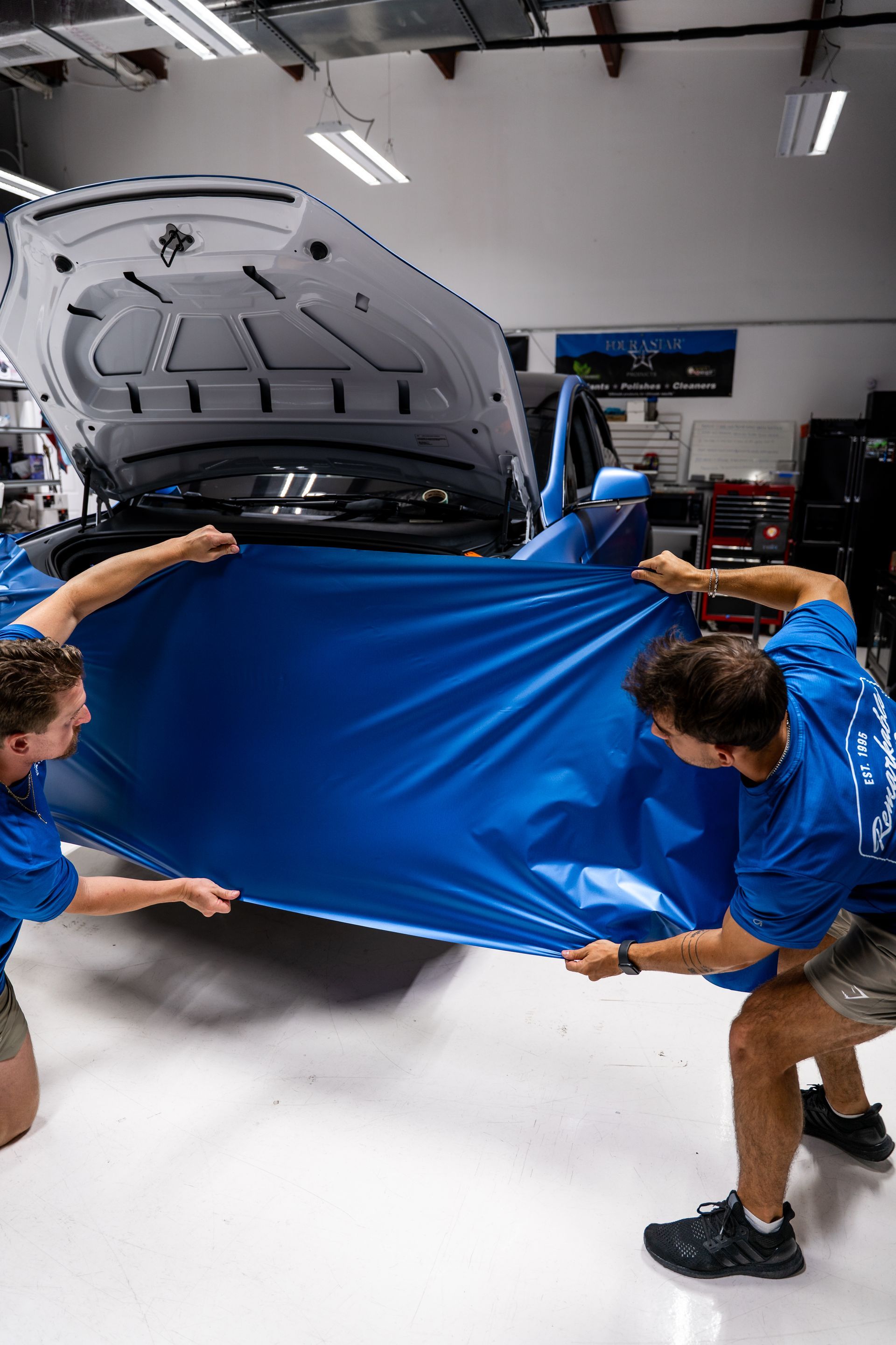 People in a garage lifting a blue cover off a car with its hood open