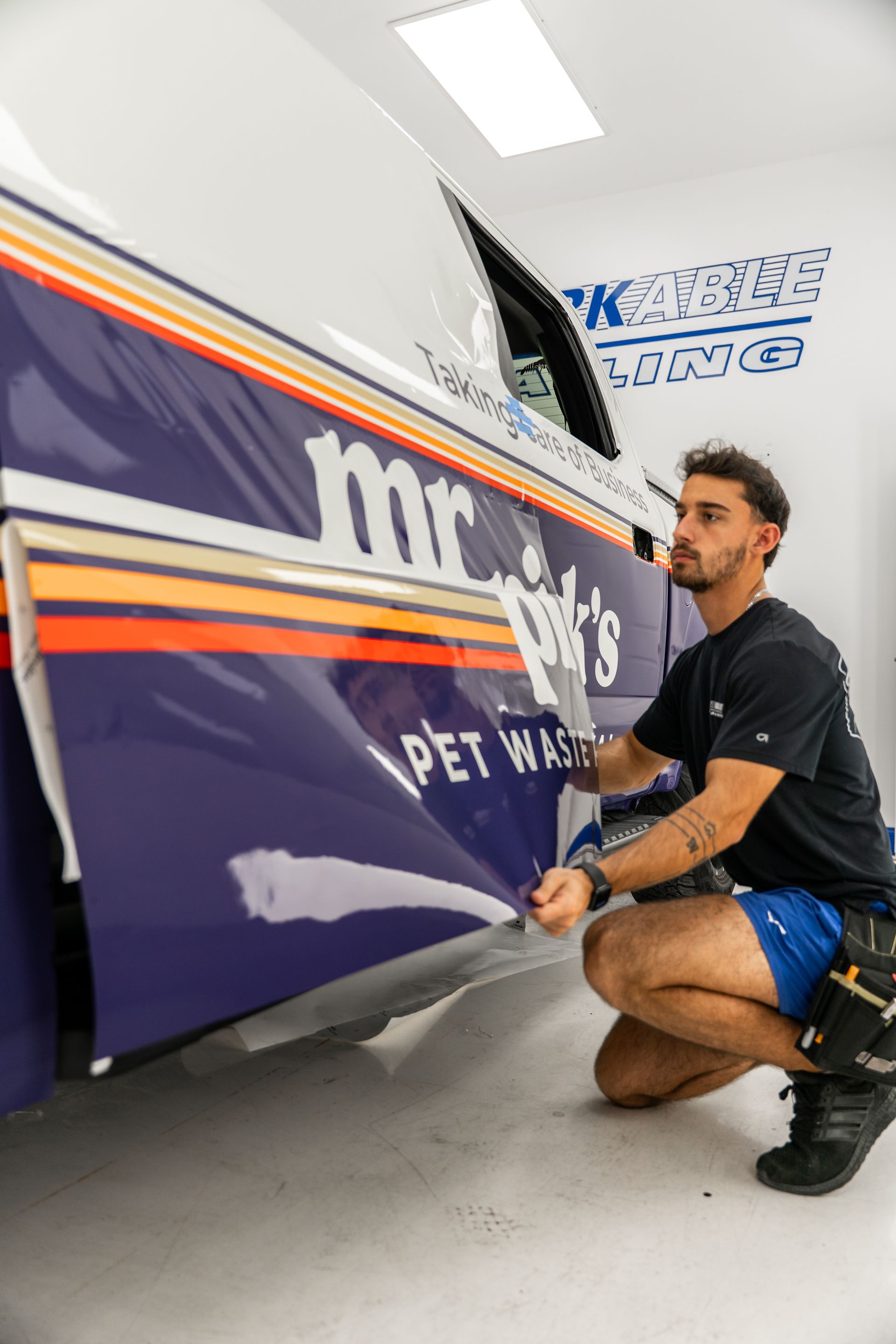 Mechanic crouches beside a purple race car in a garage, applying a decal on the side panel