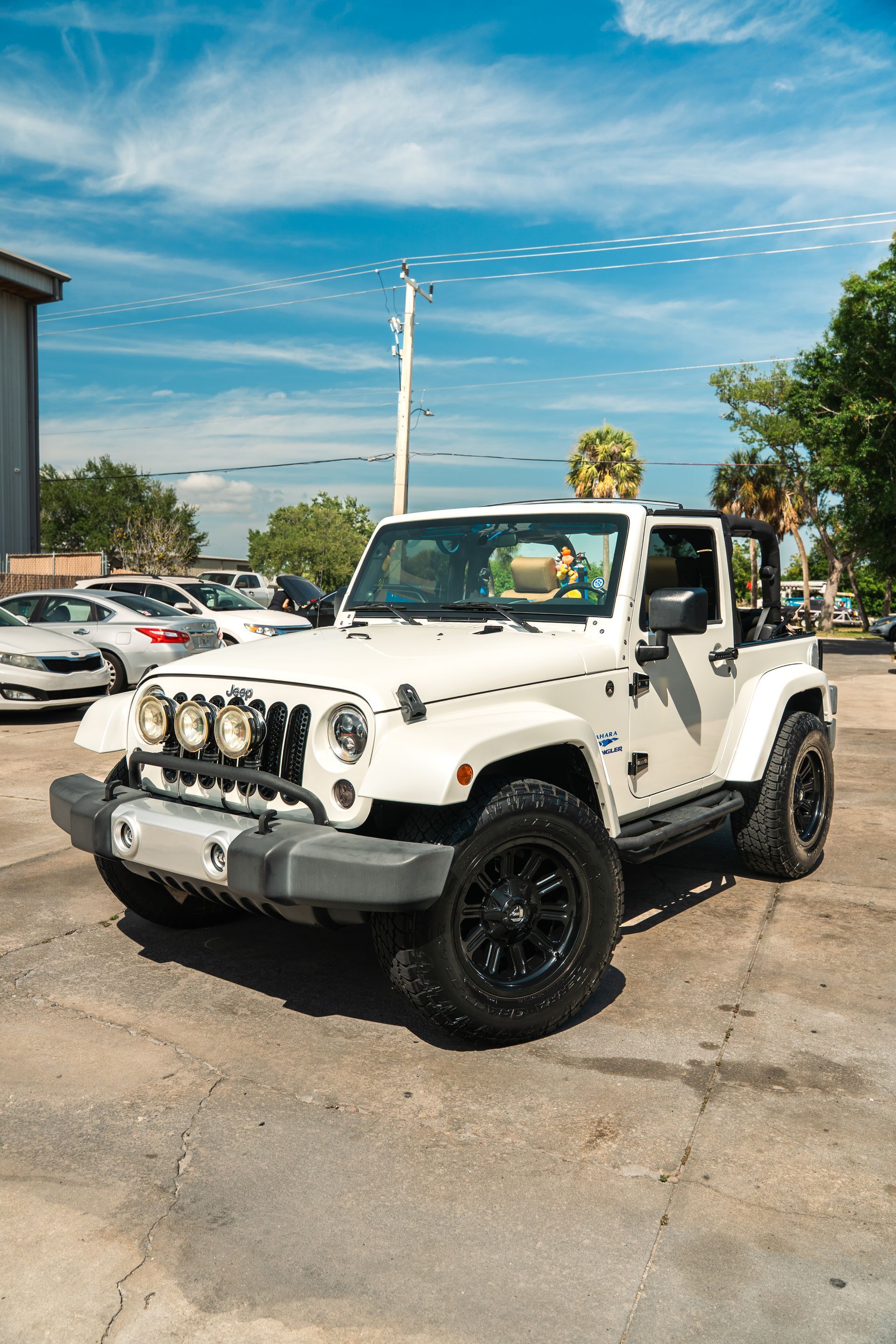 White Jeep Wrangler with black wheels parked in a lot under a blue sky, decorated with stickers and decals