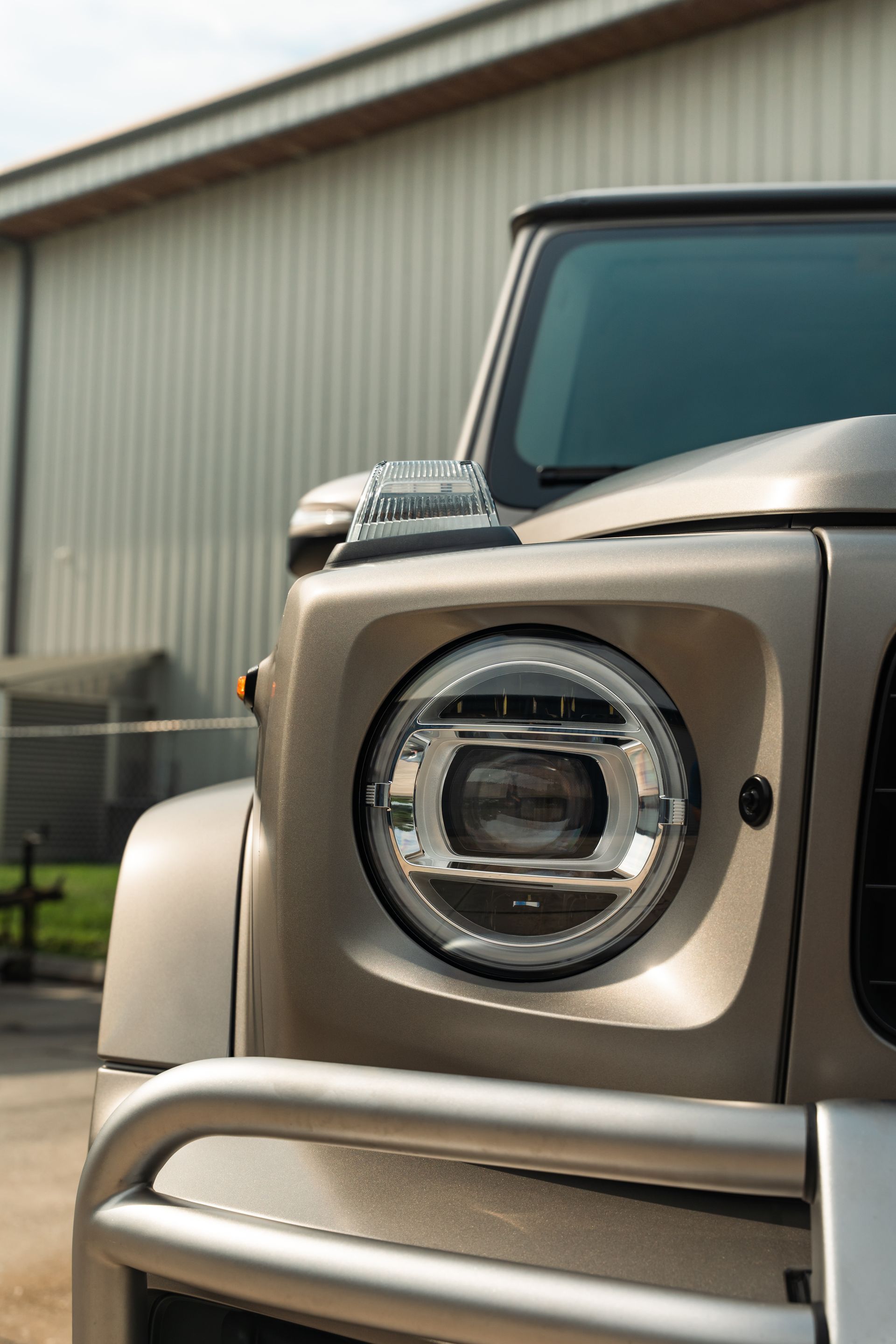 Front of a beige off-road vehicle with a round headlight, parked beside a metal building.