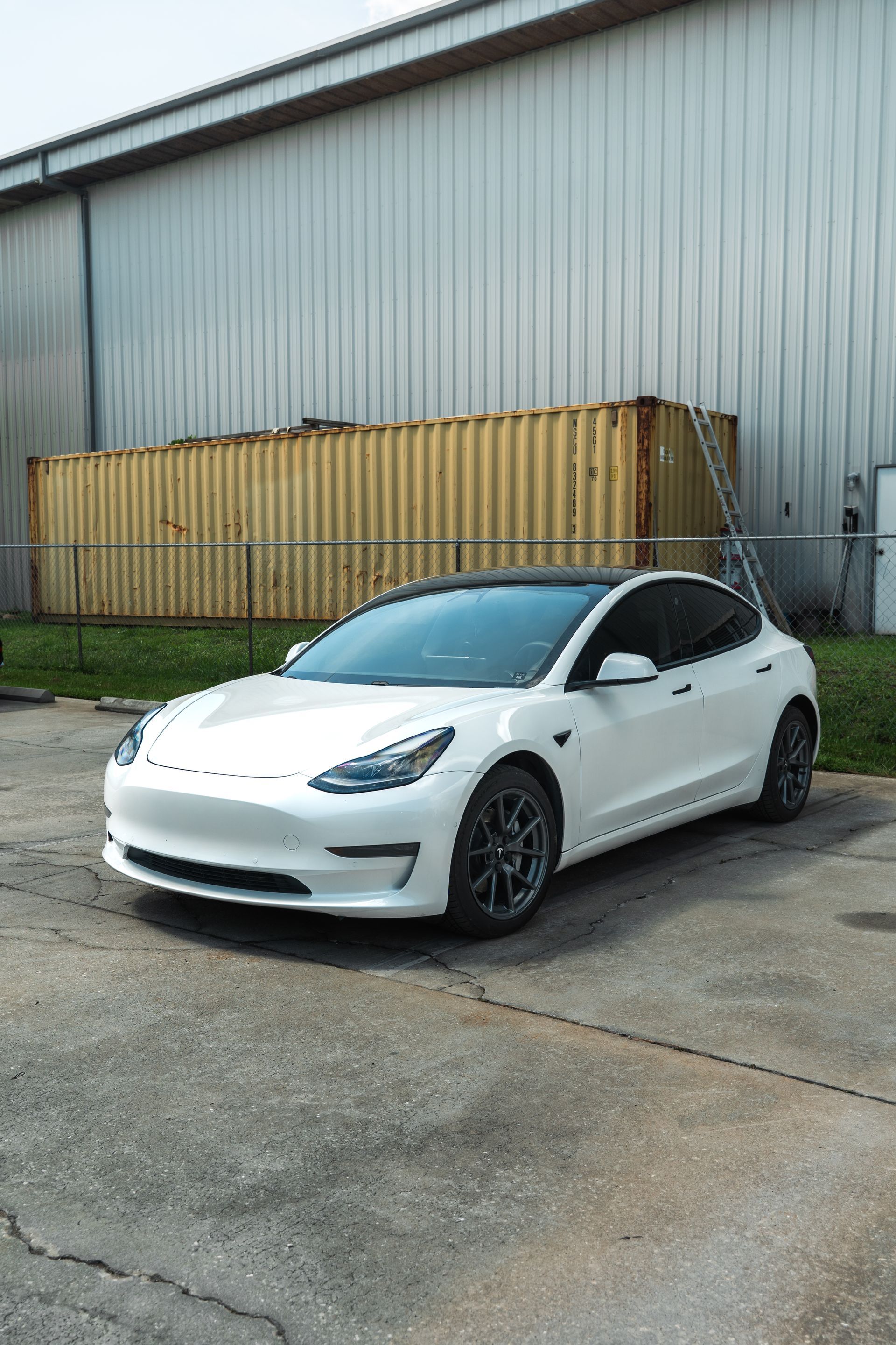White Tesla Model 3 parked beside a warehouse and shipping containers
