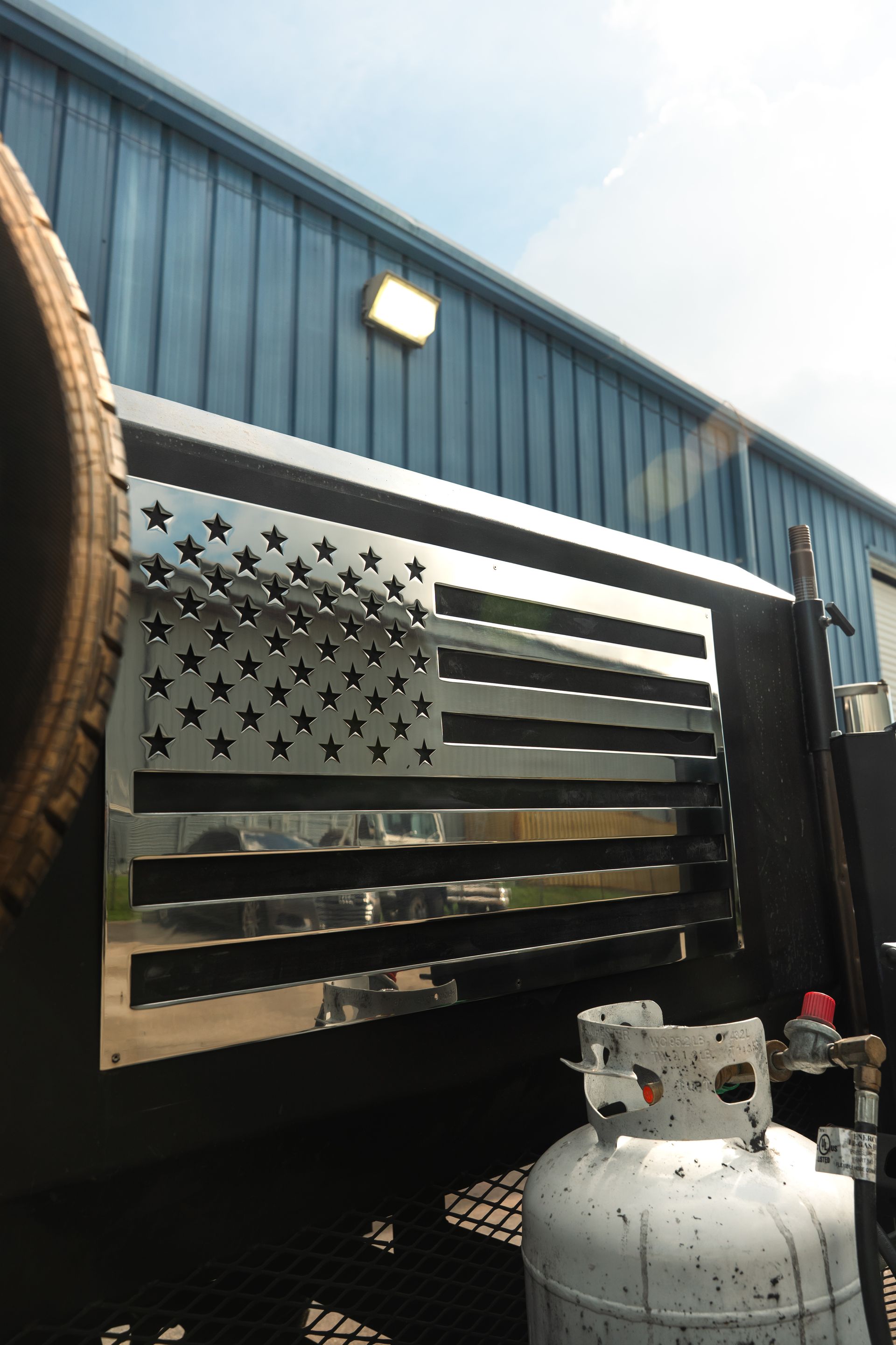 American flag metal wall art on a black trailer beside a blue building, with a propane tank in front