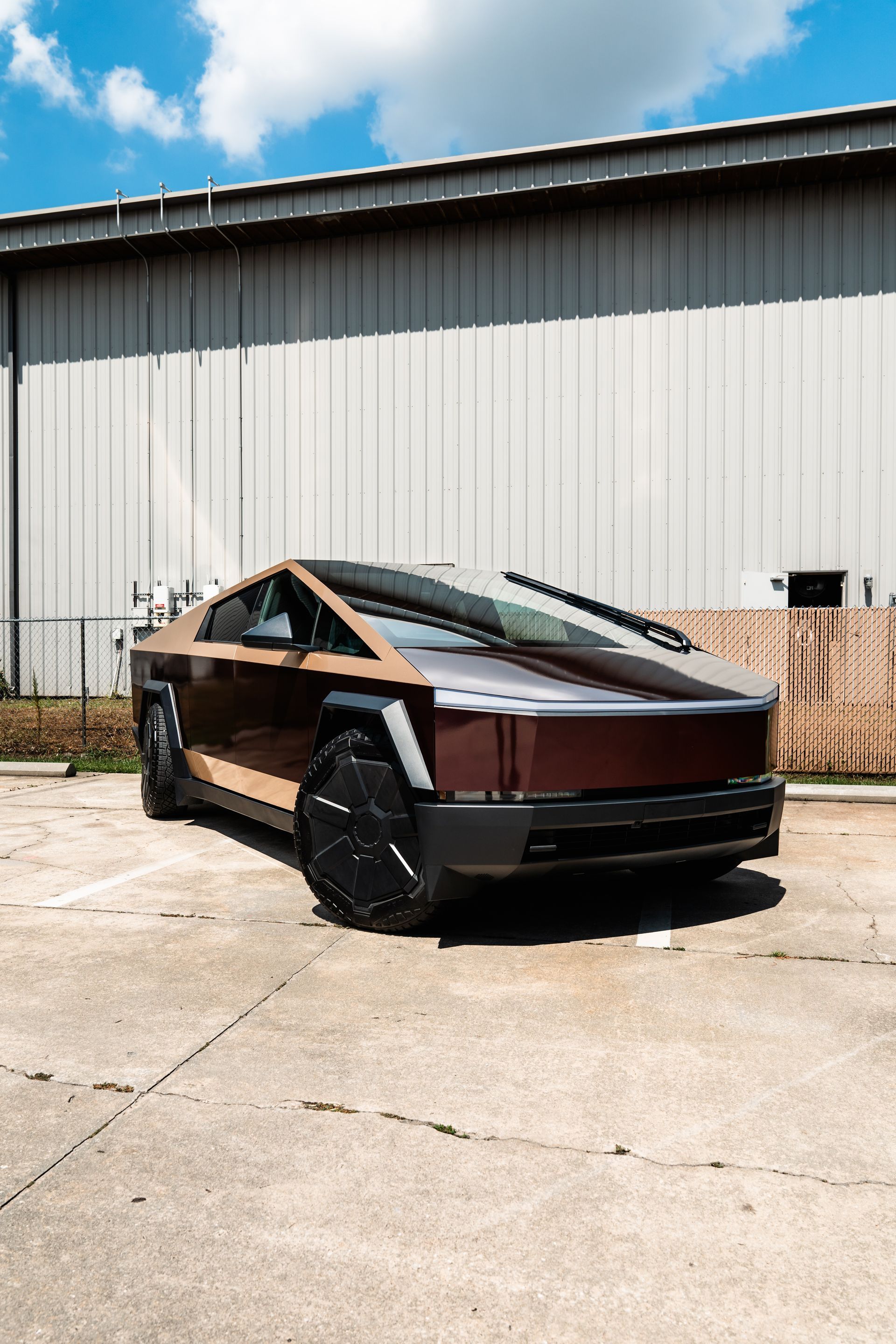 Angular brown and black futuristic car parked beside a warehouse under a bright blue sky