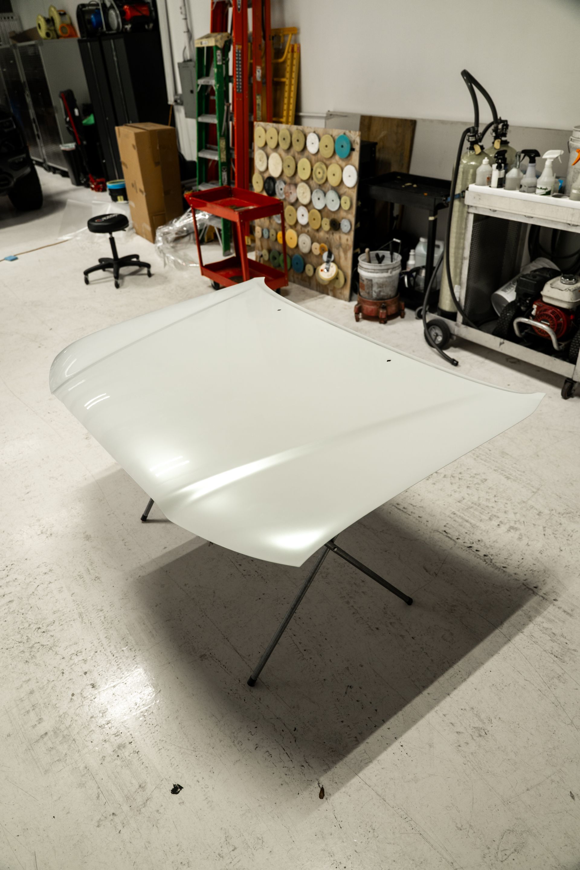White car hood on a stand in a workshop garage, with tools and equipment in the background