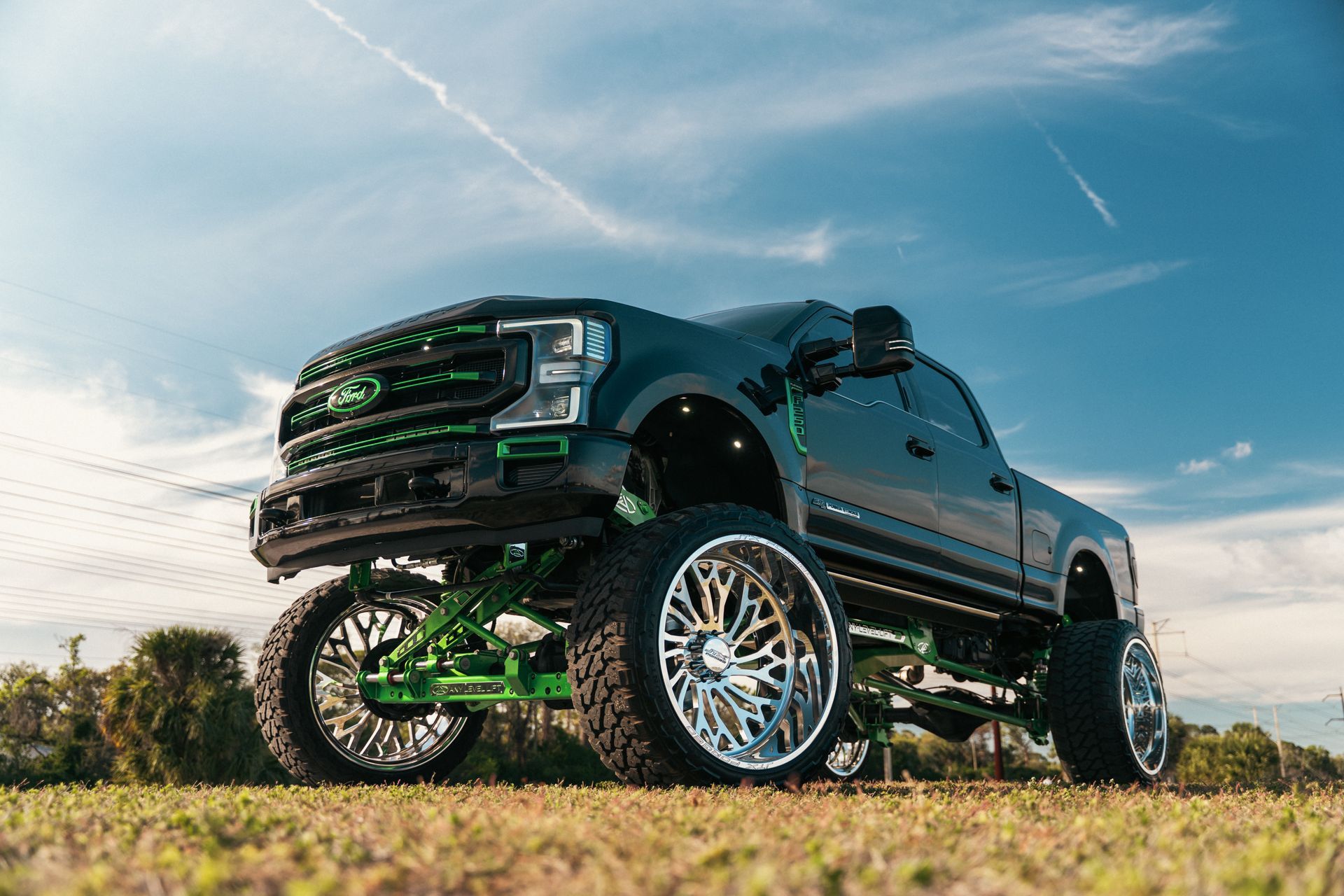Lifted black pickup truck with green suspension and chrome wheels parked in a grassy field under a blue sky