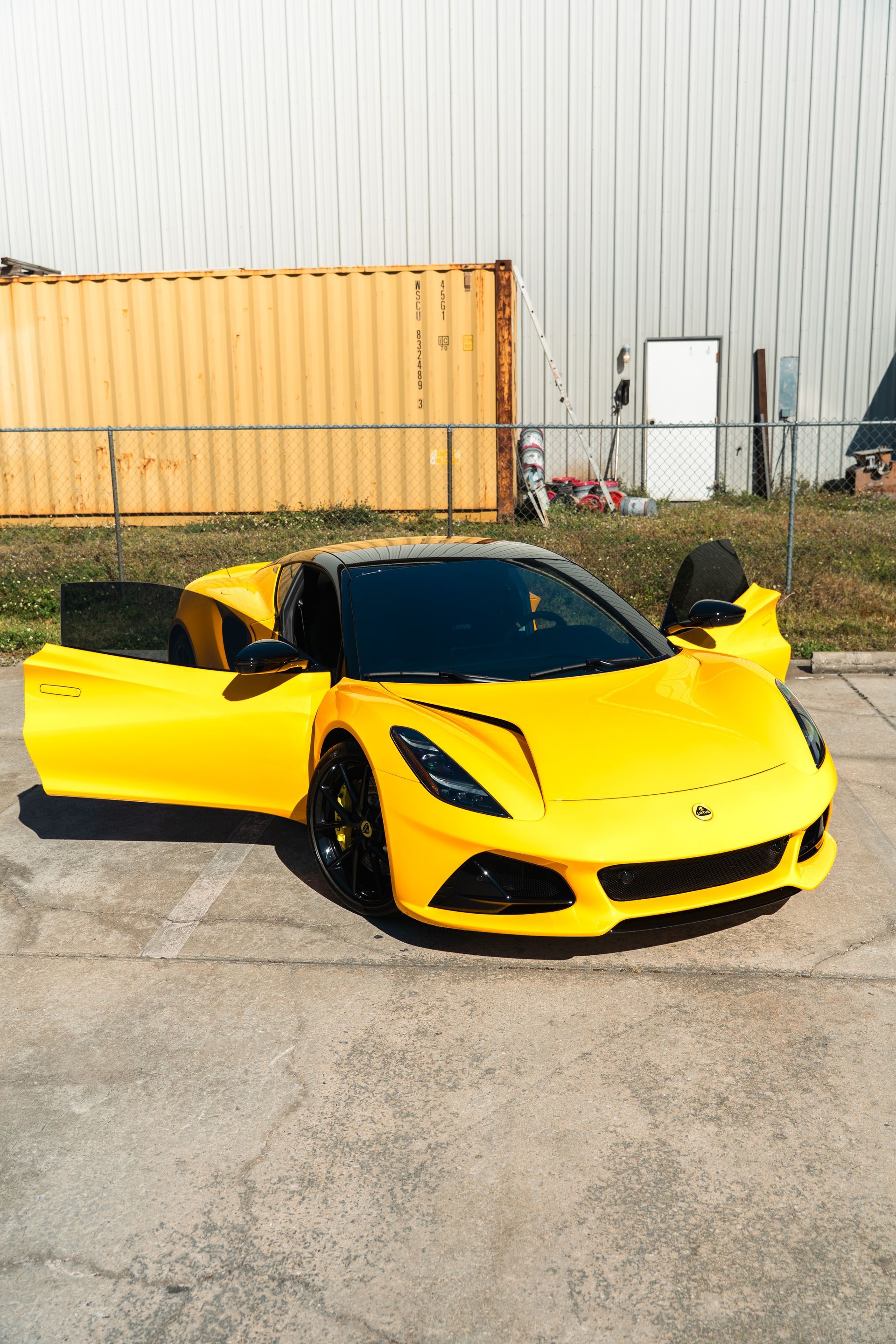 Yellow sports car with doors open, parked on pavement beside a shipping container.