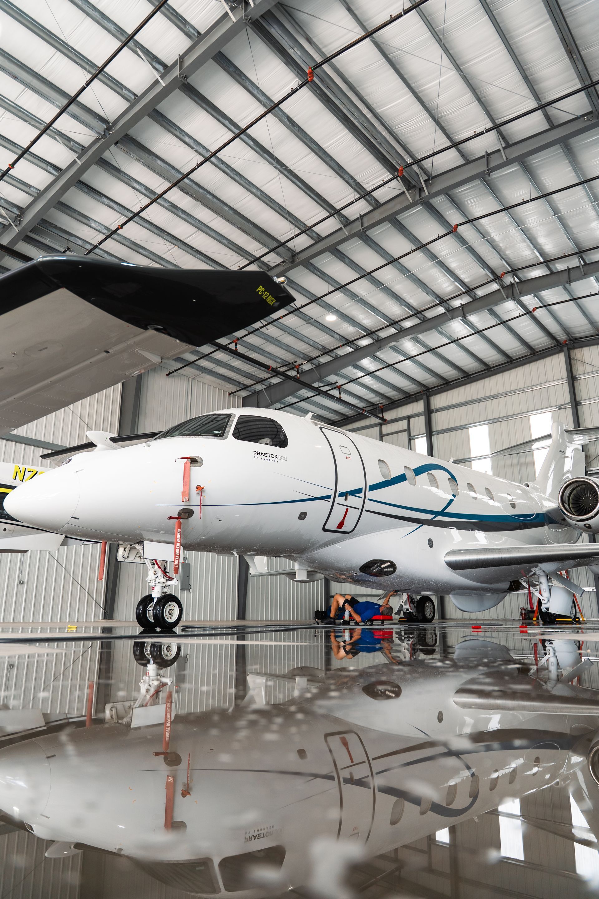 Small jet in a hangar, with a glossy floor reflecting the aircraft and curved roof beams.