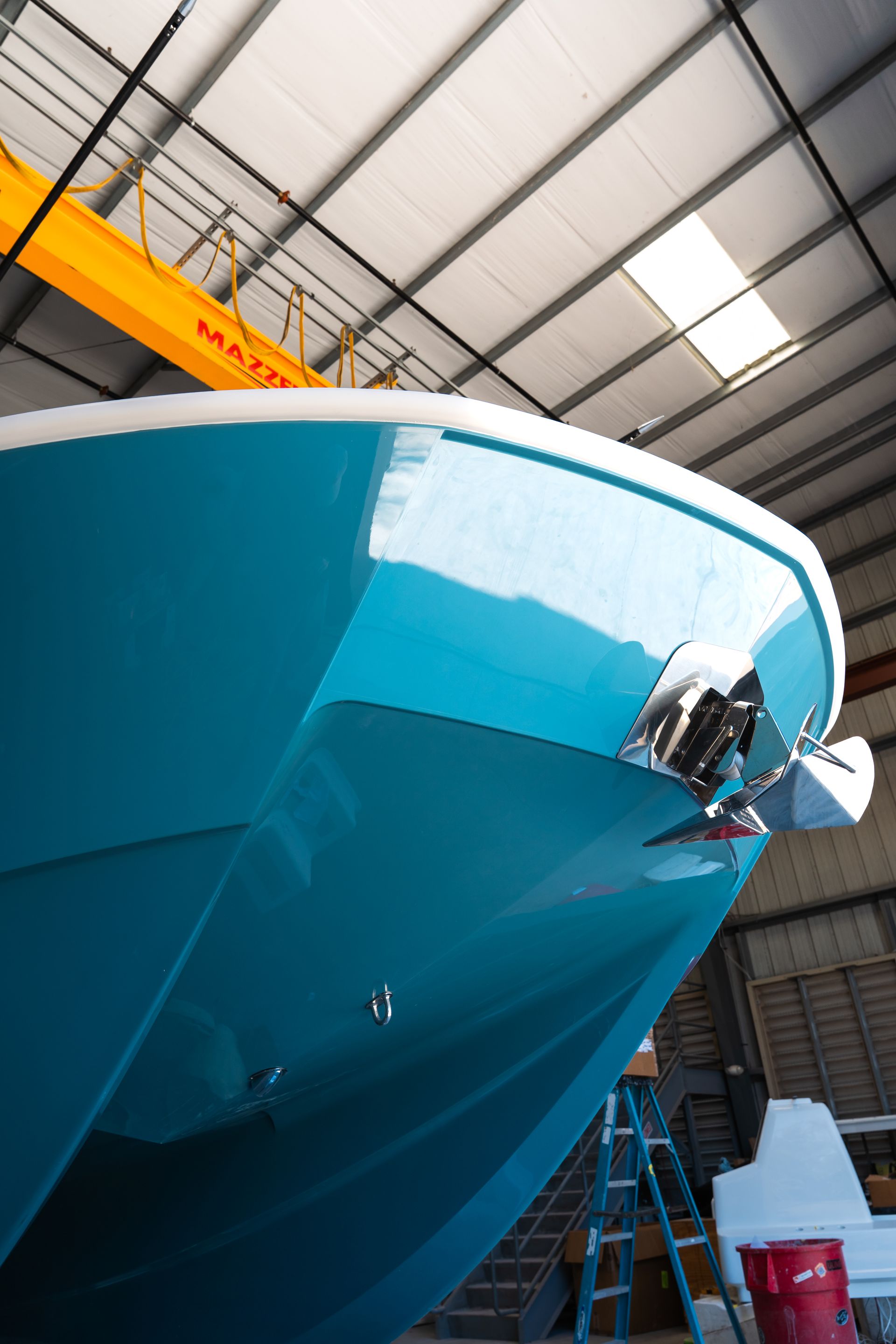 Close-up of a turquoise boat hull in an indoor shipyard with overhead beams and a ladder.