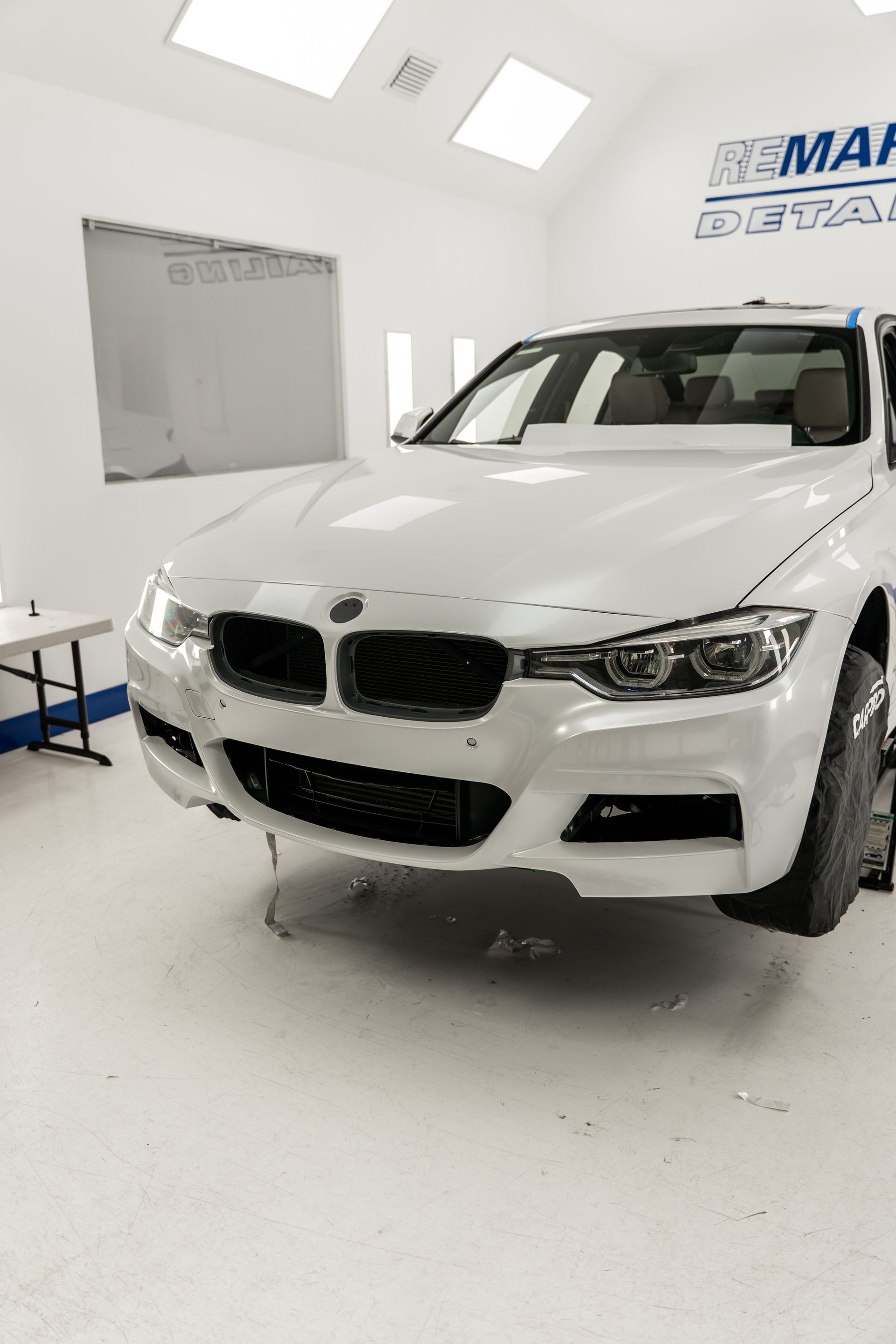 White BMW SUV in a bright auto shop, front view with hood open and windshield protected by plastic.