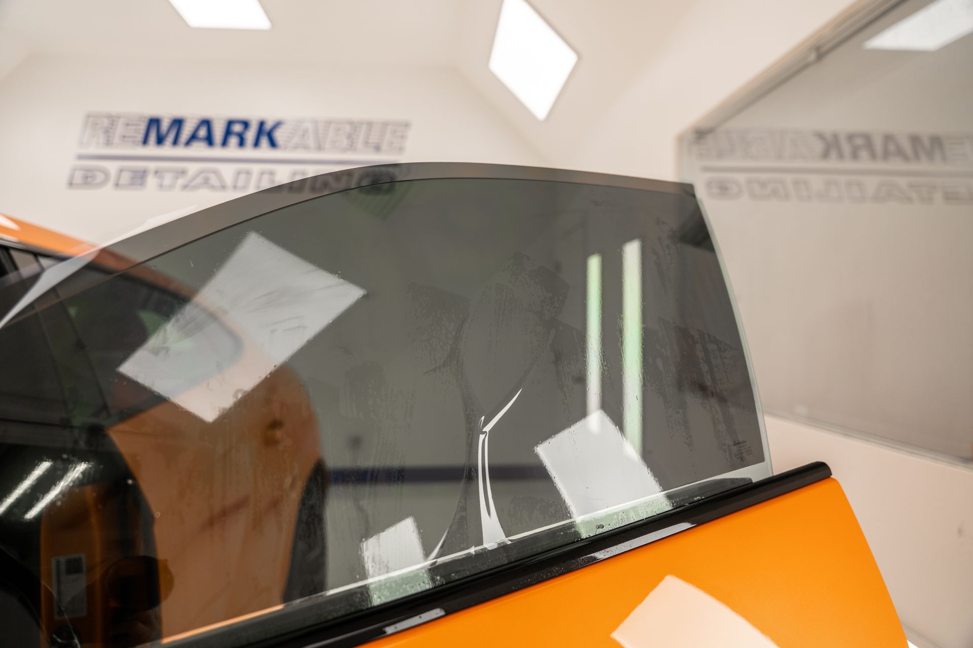 Orange car with a dark windshield in an indoor showroom, reflecting ceiling lights.