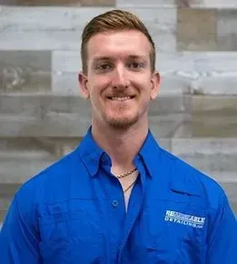 Smiling man in a blue collared work shirt against a gray stone wall