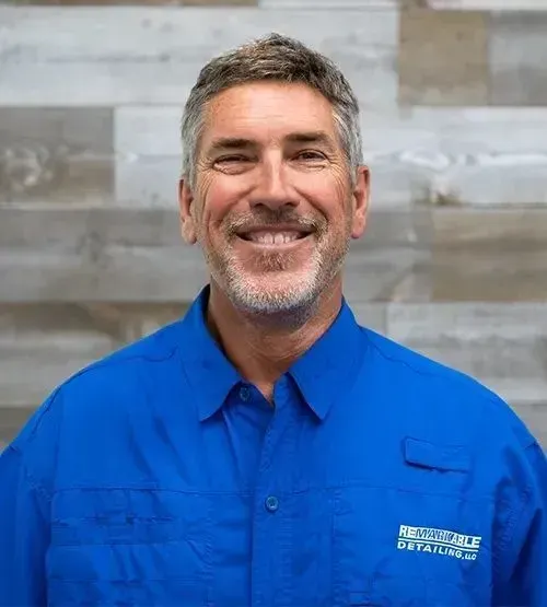 Smiling man in a bright blue button-up shirt standing against a gray wood wall.