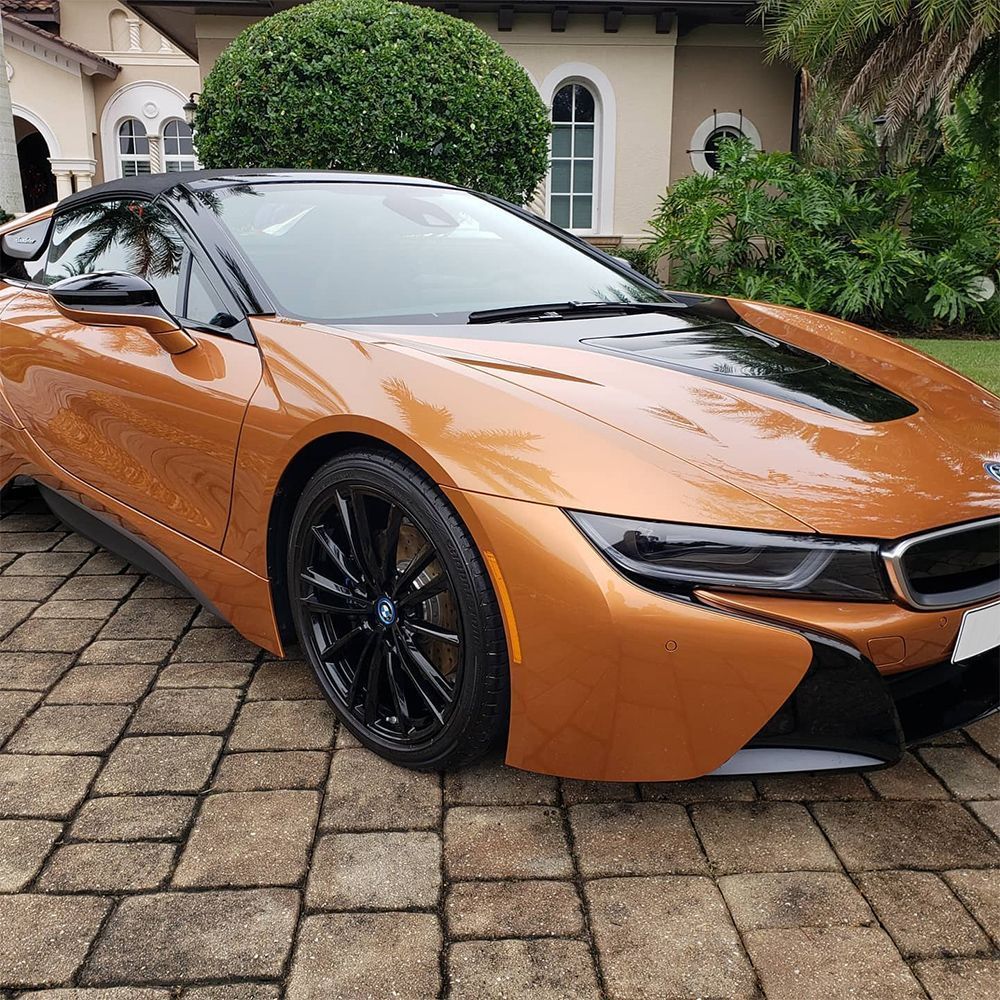 Orange sports car parked on a stone driveway outside a house, front three-quarter view.