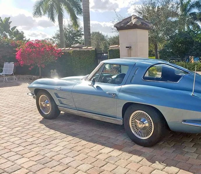 Blue vintage sports car parked on a sunlit cobblestone driveway with palm trees and flowers nearby