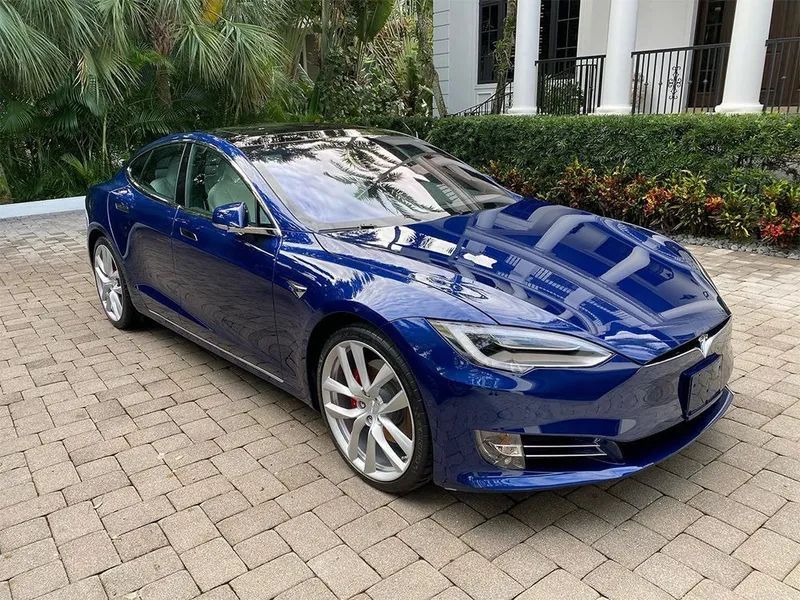 Blue Tesla sedan parked on a paved driveway in front of a building with greenery