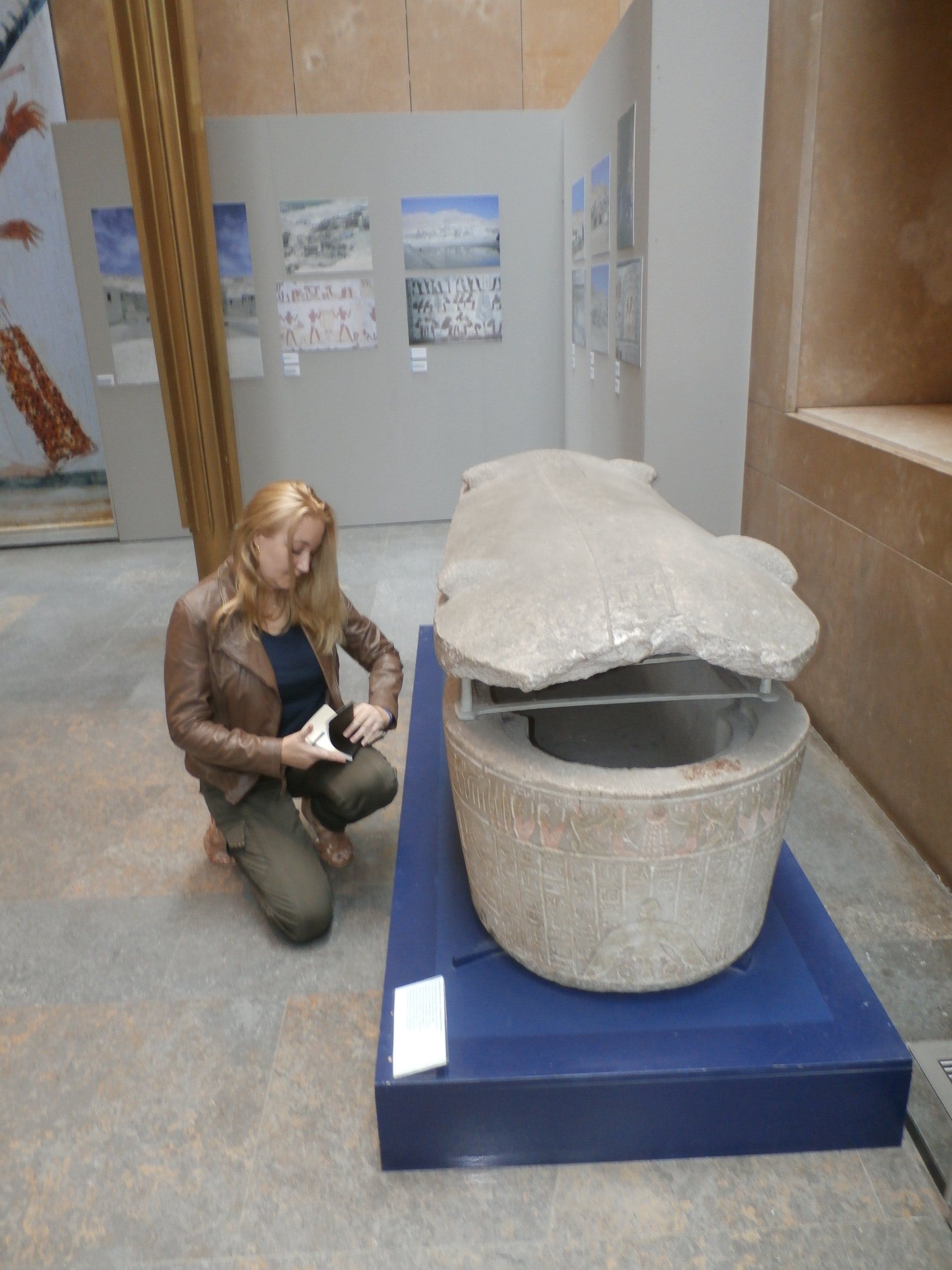 Vrouw knielt naast een groot stenen object op een blauwe sokkel in een museumtentoonstelling.