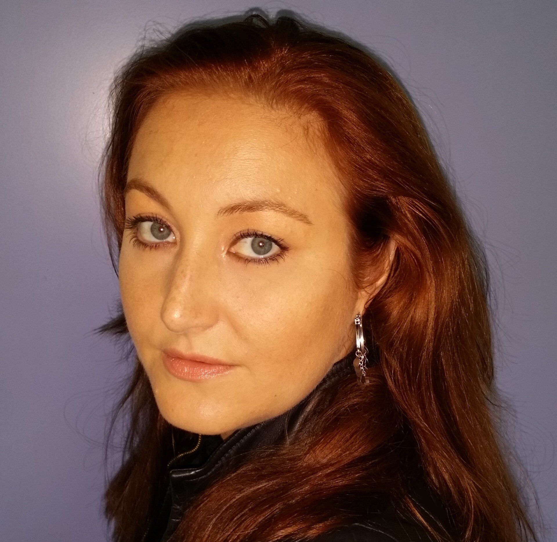 Portret van een vrouw met kastanjebruin haar en blauwe ogen tegen een zachtpaarse achtergrond.