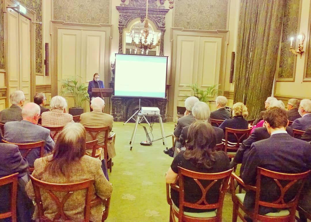 Het publiek zit in een rijkversierde zaal en kijkt naar een presentatie op een projectiescherm.