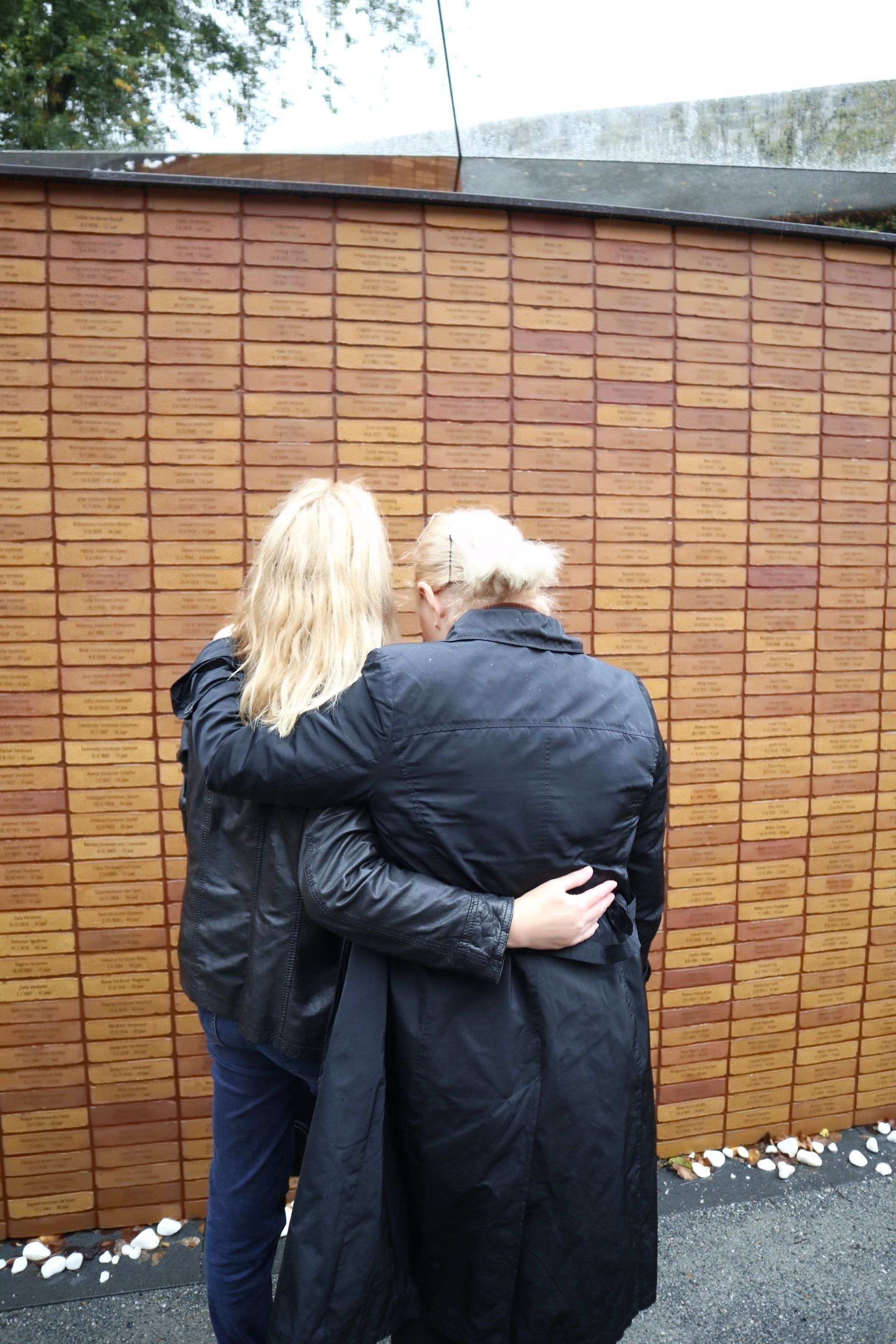 Twee mensen in donkere jassen staan ​​arm in arm voor een houten muur buiten.