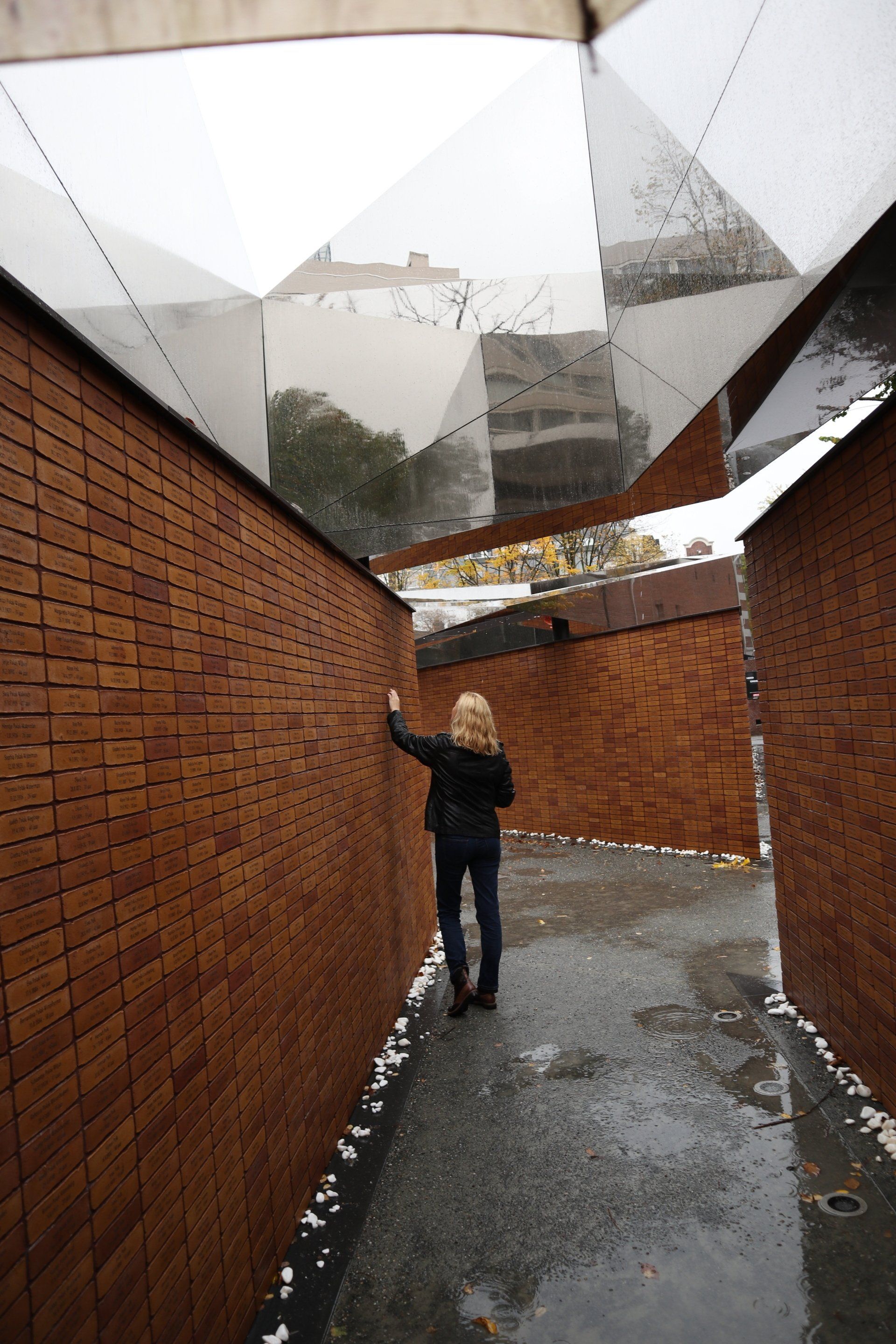 Een persoon staat in een smalle bakstenen steeg en kijkt omhoog naar de geometrische, spiegelende panelen boven zijn hoofd.